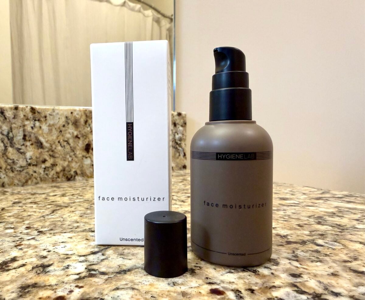 The photo features an unscented "face moisturizer" from the brand Hygiene Lab, shown as a matte-brown pump bottle next to its white packaging and a black cap on a granite countertop.