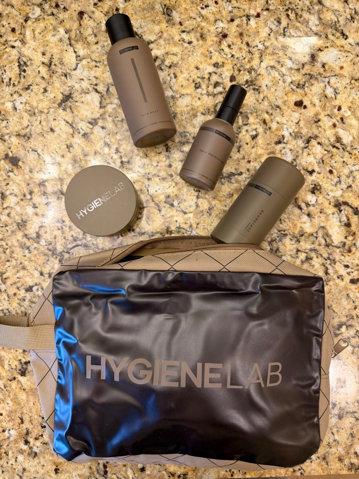 The photo displays an assortment of brown Hygiene Lab personal care products, including face wash, moisturizer, deodorant, and a styling gel jar, arranged above a tan and black quilted toiletry bag on a granite countertop.