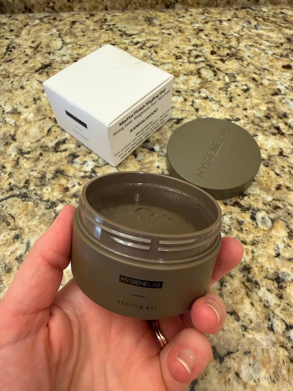 The photo shows a person's hand holding an open jar of sandalwood-scented "styling gel" by Hygiene Lab, revealing the product inside, next to its white box and lid on a granite countertop.
