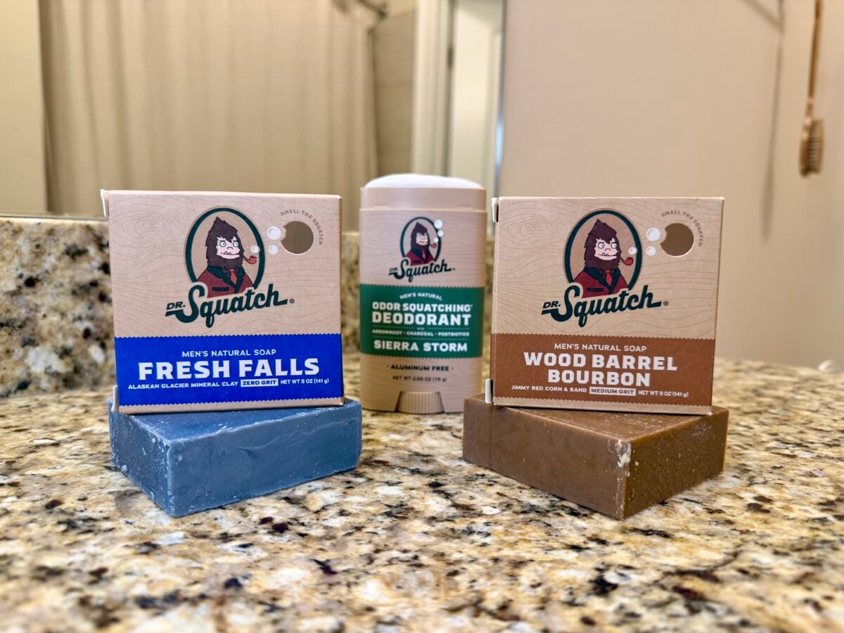 Dr. Squatch Fresh Falls and Wood Barrel Bourbon soap boxes displayed on unwrapped bars with a Sierra Storm aluminum-free deodorant on a bathroom countertop.
