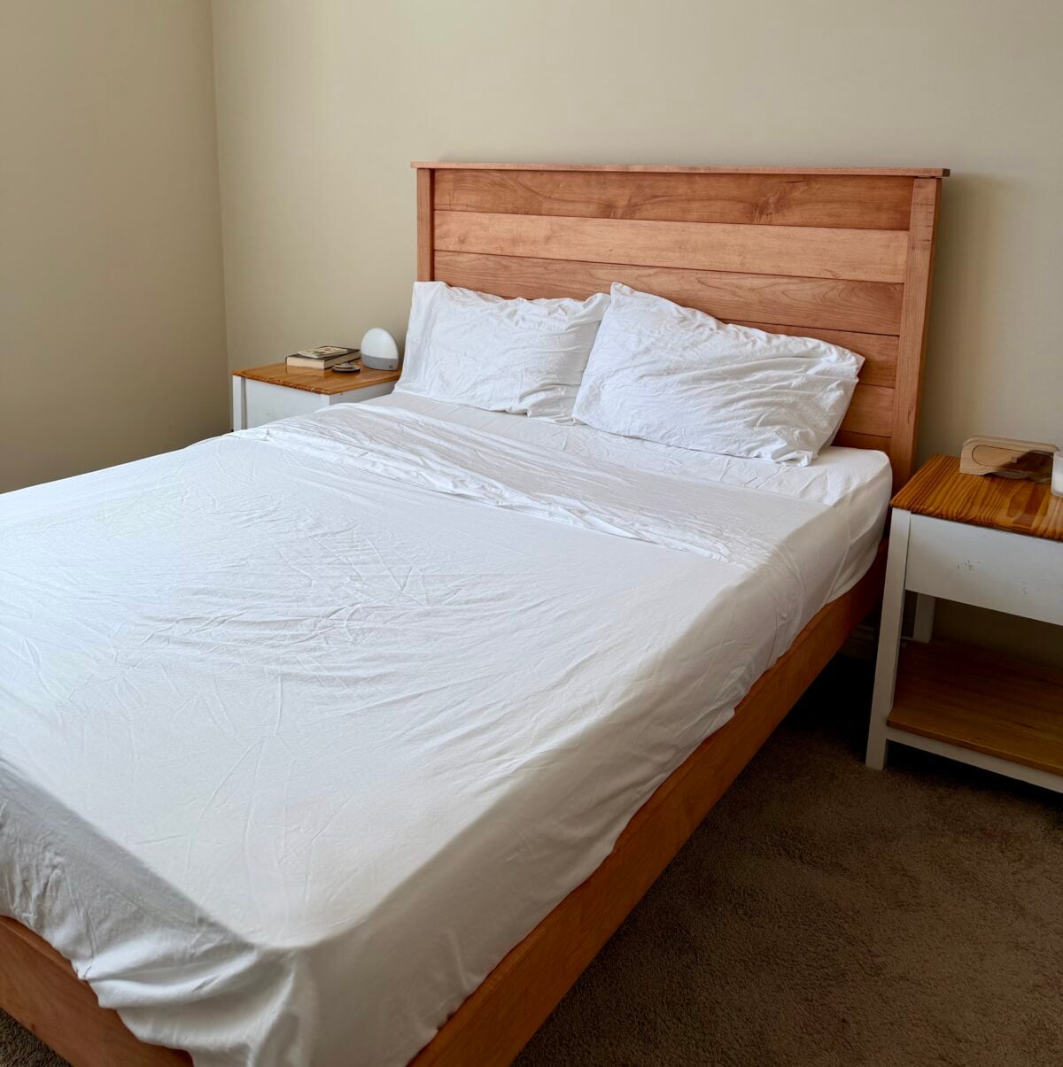A neatly made bed with a wooden headboard and white bedding is centered between two white nightstands. One nightstand has books and a lamp, while the other has a wooden surface. The room has beige walls and carpeted flooring.