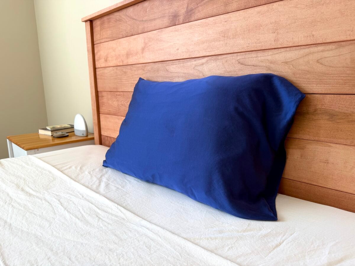 A wooden headboard and white bed with a single blue pillow. A nightstand with a lamp and a small stack of books is visible next to the bed.