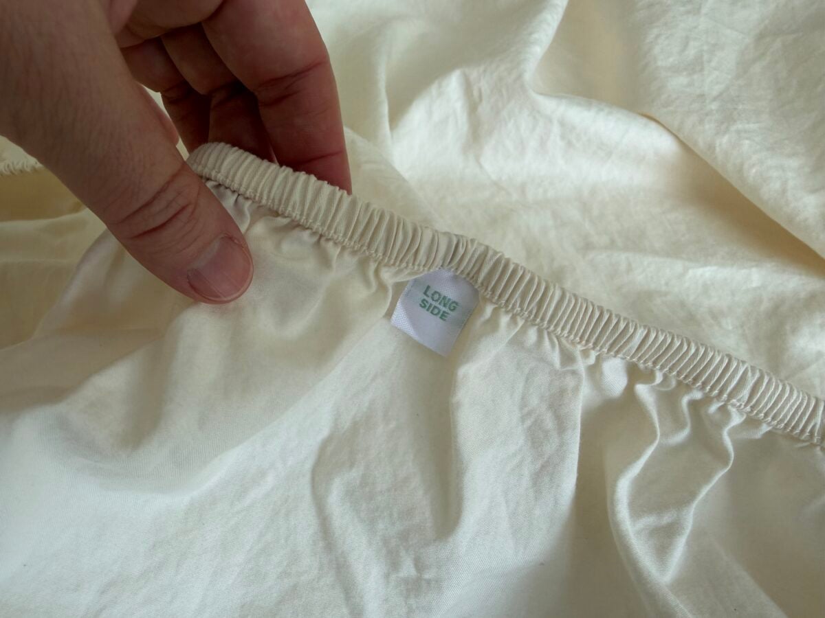A hand holds the elastic edge of a cream-colored fitted sheet, revealing a sewn-in white tag labeled LONG SIDE in green text. The sheet is spread out on a flat surface.