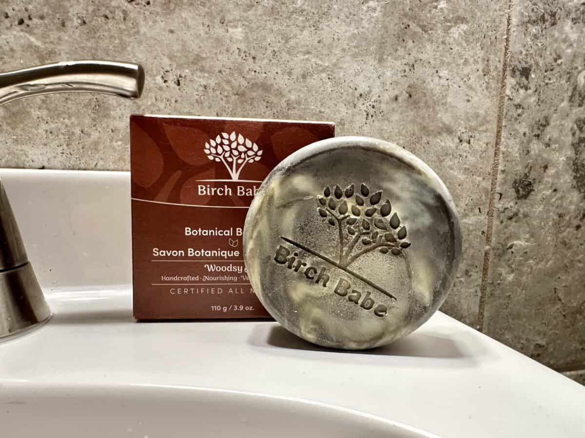 The photo shows a round, marbled Birch Babe bar soap embossed with the brand logo sitting on a white bathroom sink next to its brown cardboard packaging.