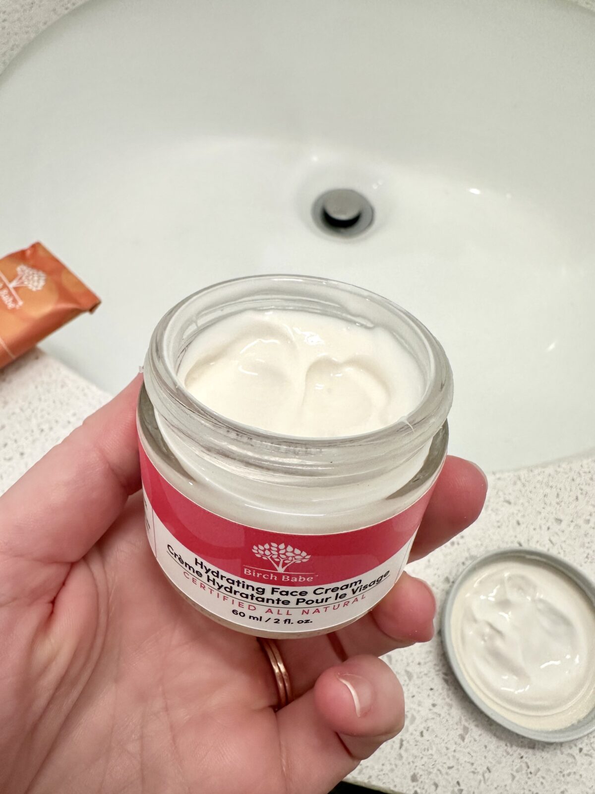 The photo shows a hand holding an open jar of Birch Babe "Hydrating Face Cream" over a white bathroom sink, revealing the thick white cream inside with the lid resting on the speckled counter nearby.
