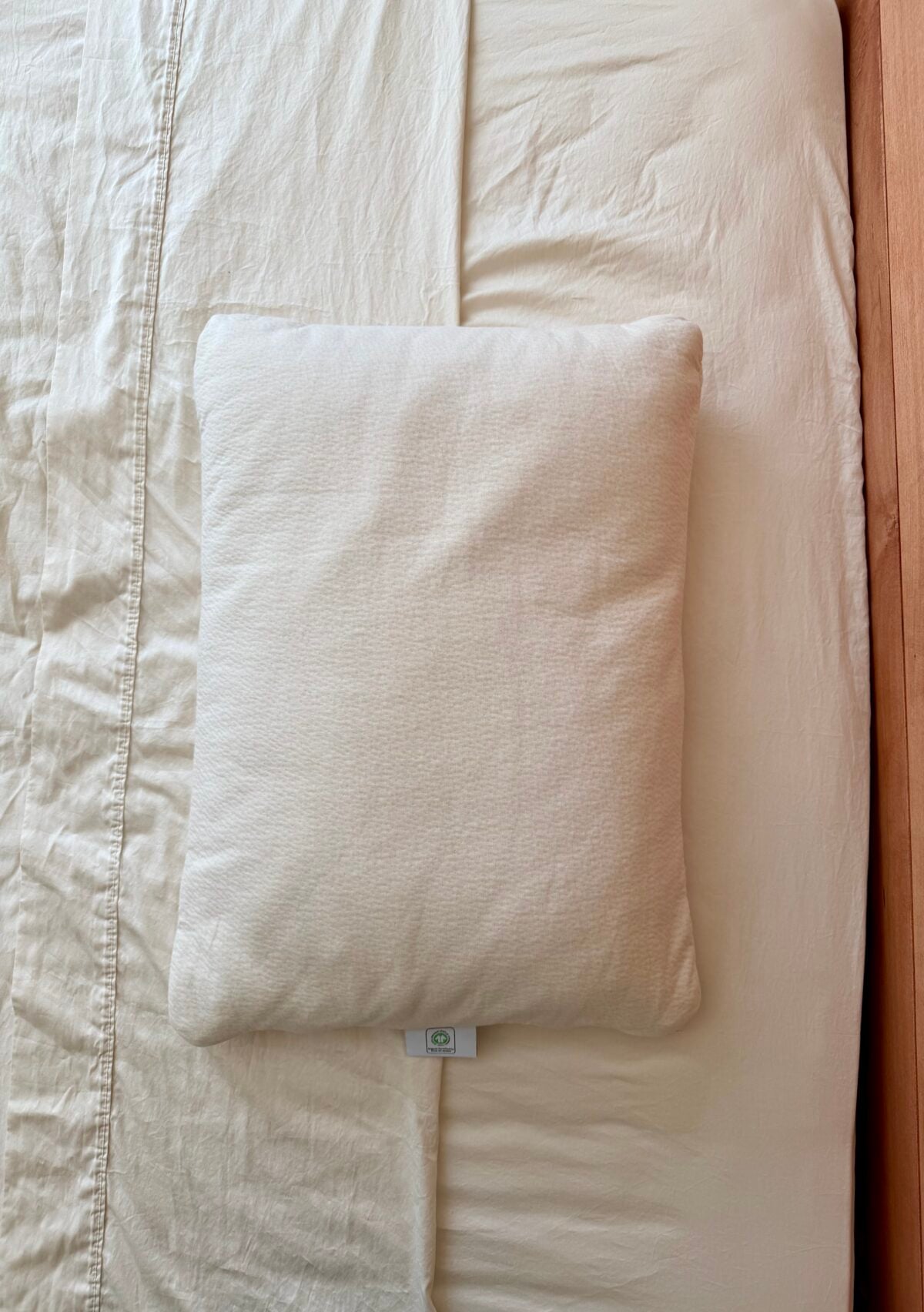 A square, cream-colored pillow is placed on a neatly made bed with matching cream sheets and a mattress visible to the right. The bed has a wooden edge on the right side.