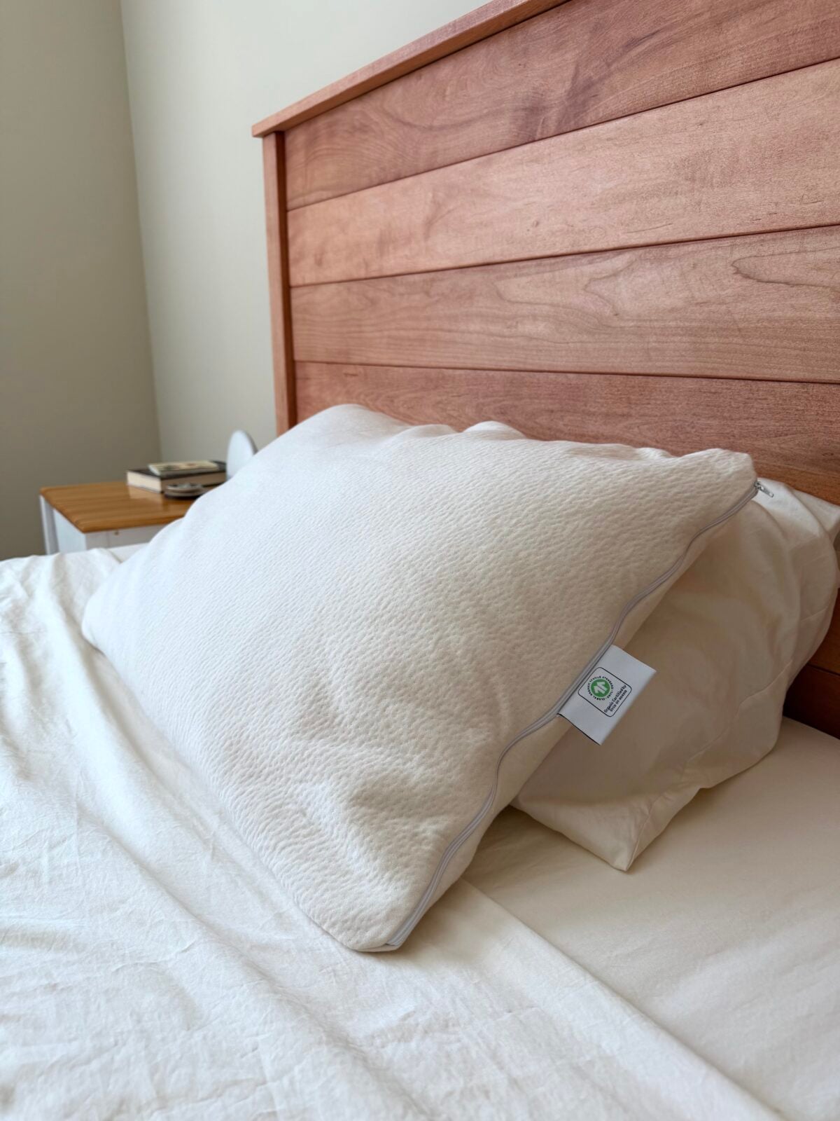 A close-up of a neatly made bed with a large white pillow and a wooden headboard. A small bedside table with a few items, including a book, is partially visible in the background.