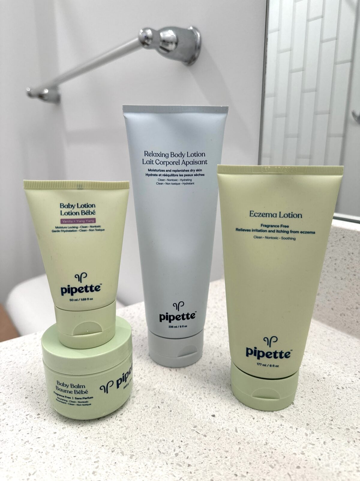 The photo displays an assortment of Pipette brand skincare products, including tubes of Baby Lotion, Relaxing Body Lotion, and Eczema Lotion, along with a jar of Baby Balm, arranged on a white speckled bathroom countertop.