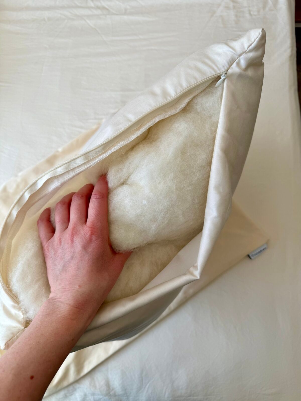 A hand is seen opening the zipper of a cream-colored pillow cover, revealing the white, fluffy fill inside. The pillow rests on a matching cream-colored surface.