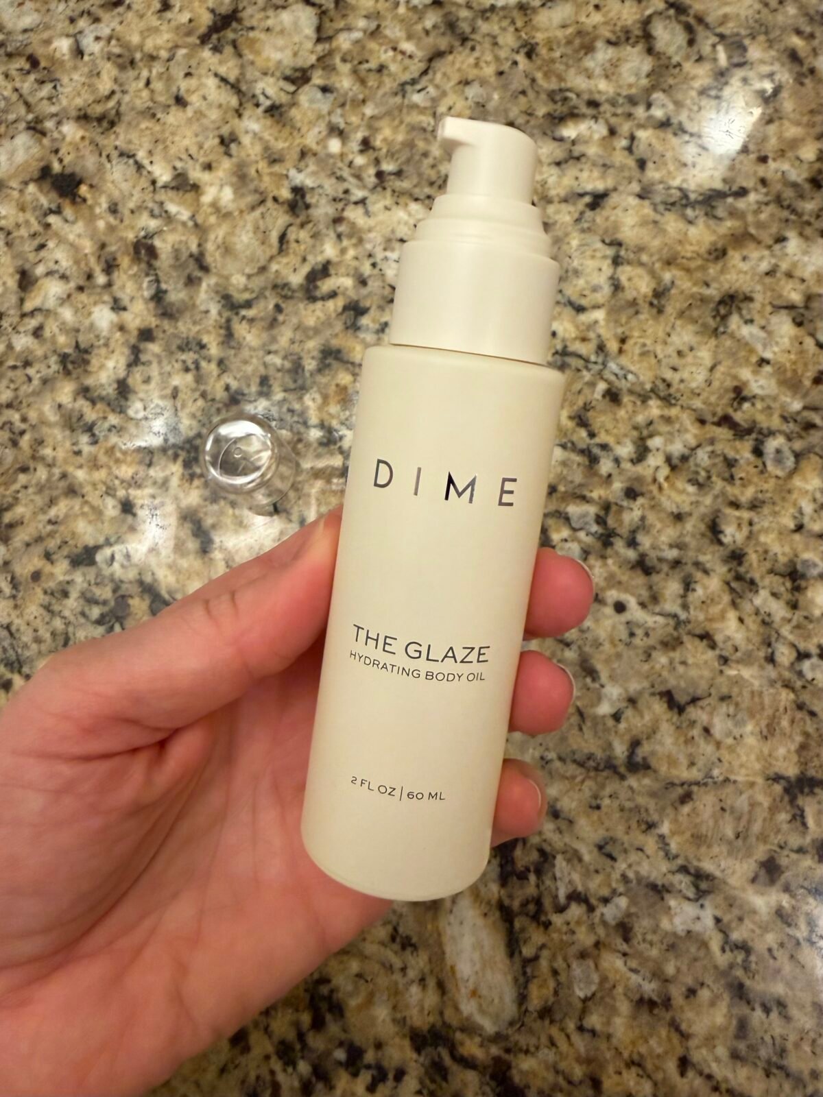 Hand holding DIME The Glaze body oil bottle showing pump dispenser and 2 fl oz size on granite counter
