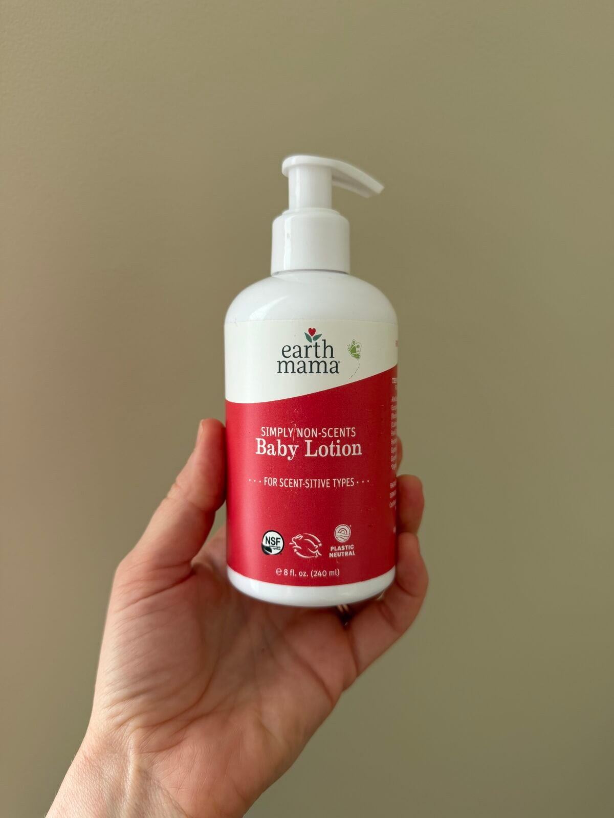 The photo shows a hand holding an 8 fl. oz. white pump bottle of Earth Mama "Simply Non-Scents Baby Lotion" against a neutral, plain wall.