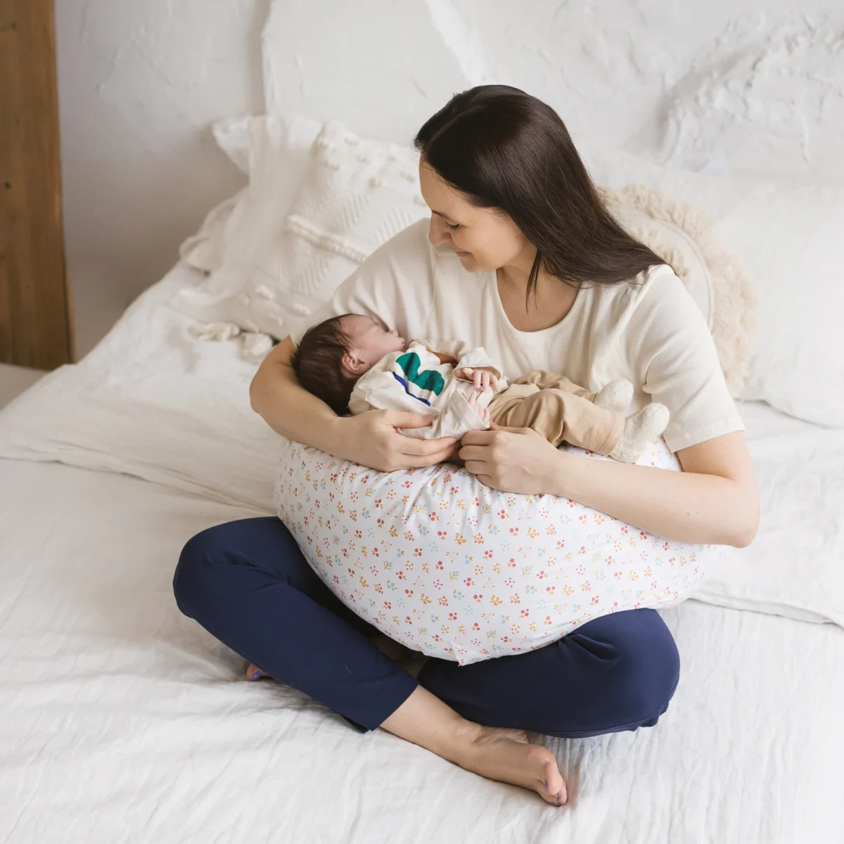 Mother sitting cross-legged on bed using a floral-patterned nursing pillow to support infant during breastfeeding