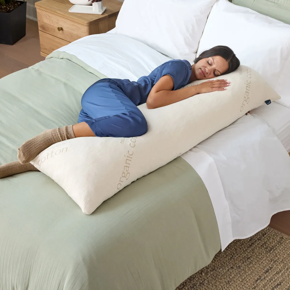 The photo shows a woman in blue pajamas sleeping on a bed while hugging a long, white organic cotton body pillow.