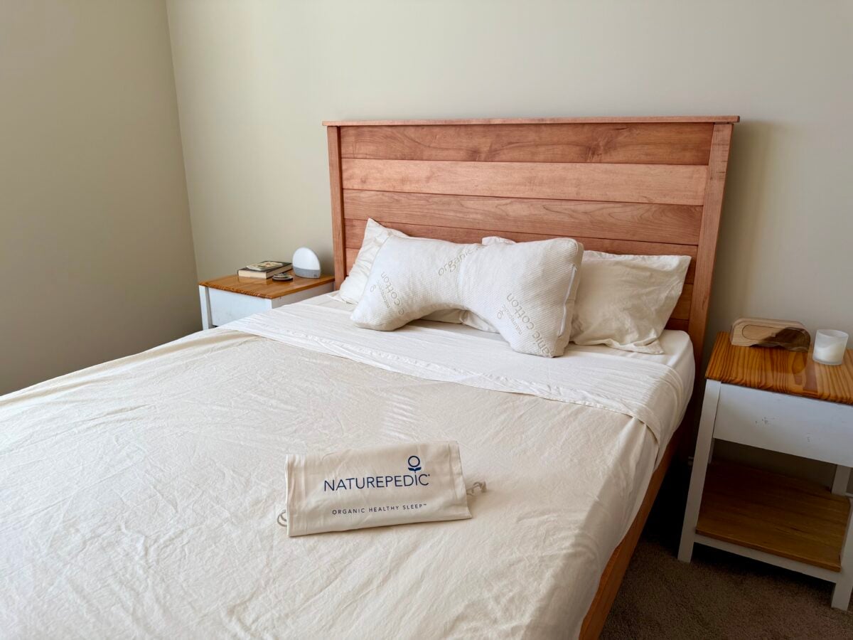 A neatly made bed with a wooden headboard, two pillows, and a white sheet. On the bed is a Naturepedic bag. There are nightstands on each side, one with a lamp and the other with a candle.