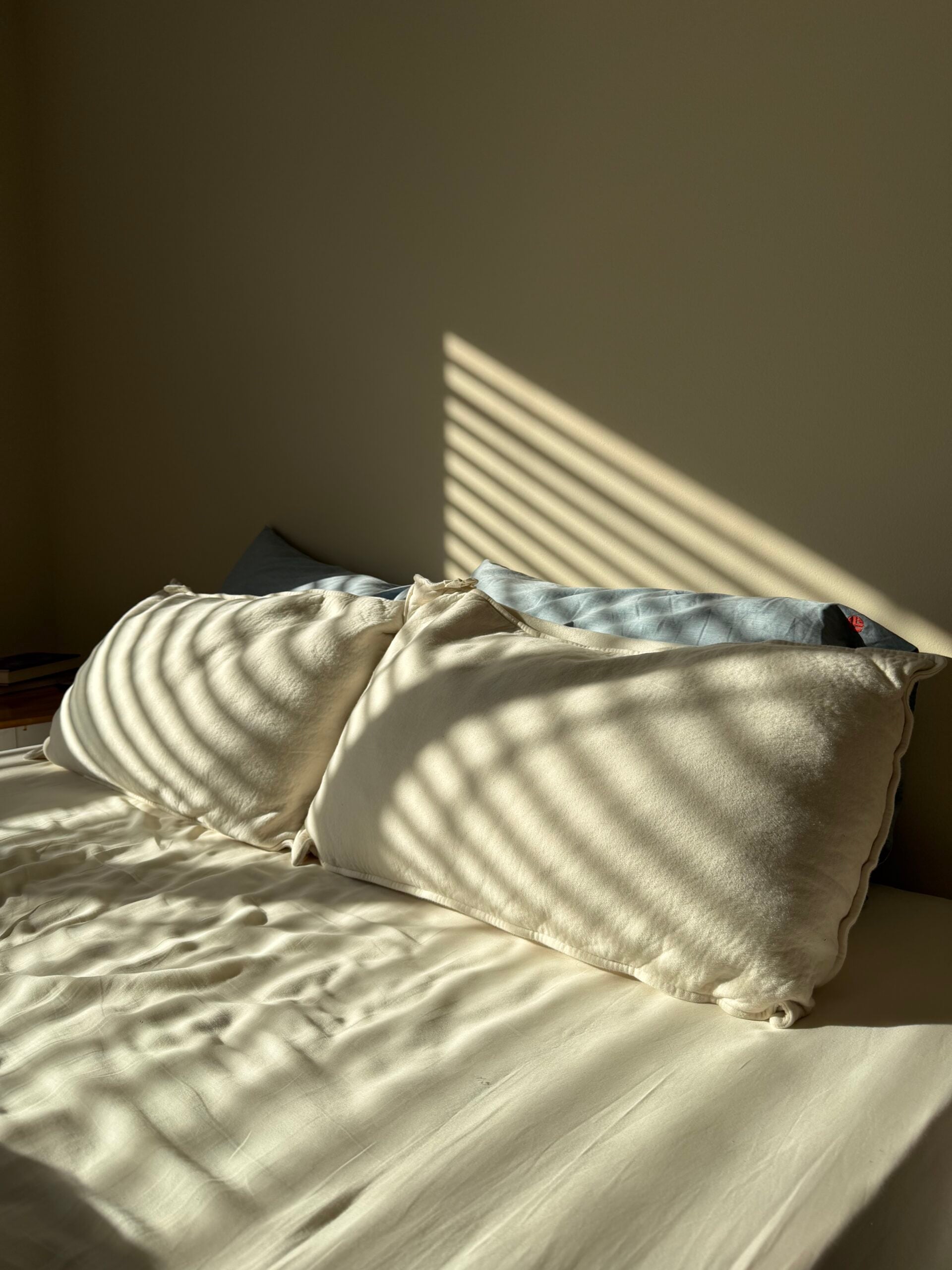 Two organic cotton pillows with natural-colored covers on a bed with sunlight streaming through window blinds