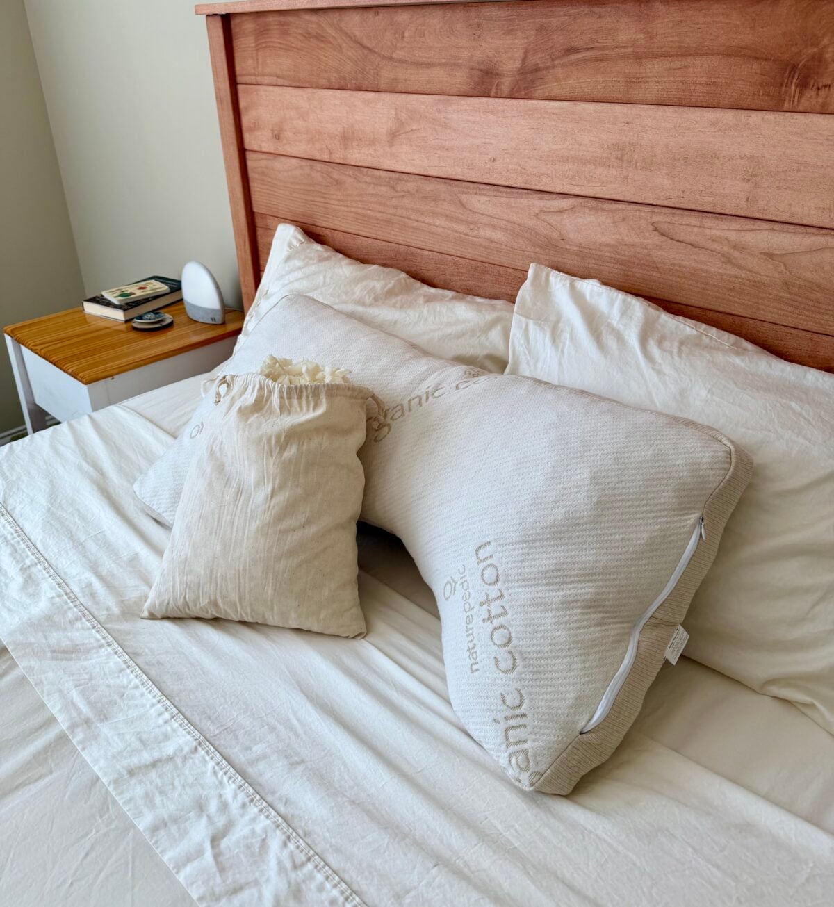 A neatly made bed with cream-colored sheets, two large pillows labeled organic cotton, and a smaller drawstring bag resting on top. A wooden headboard and a bedside table with books and a clock are visible.