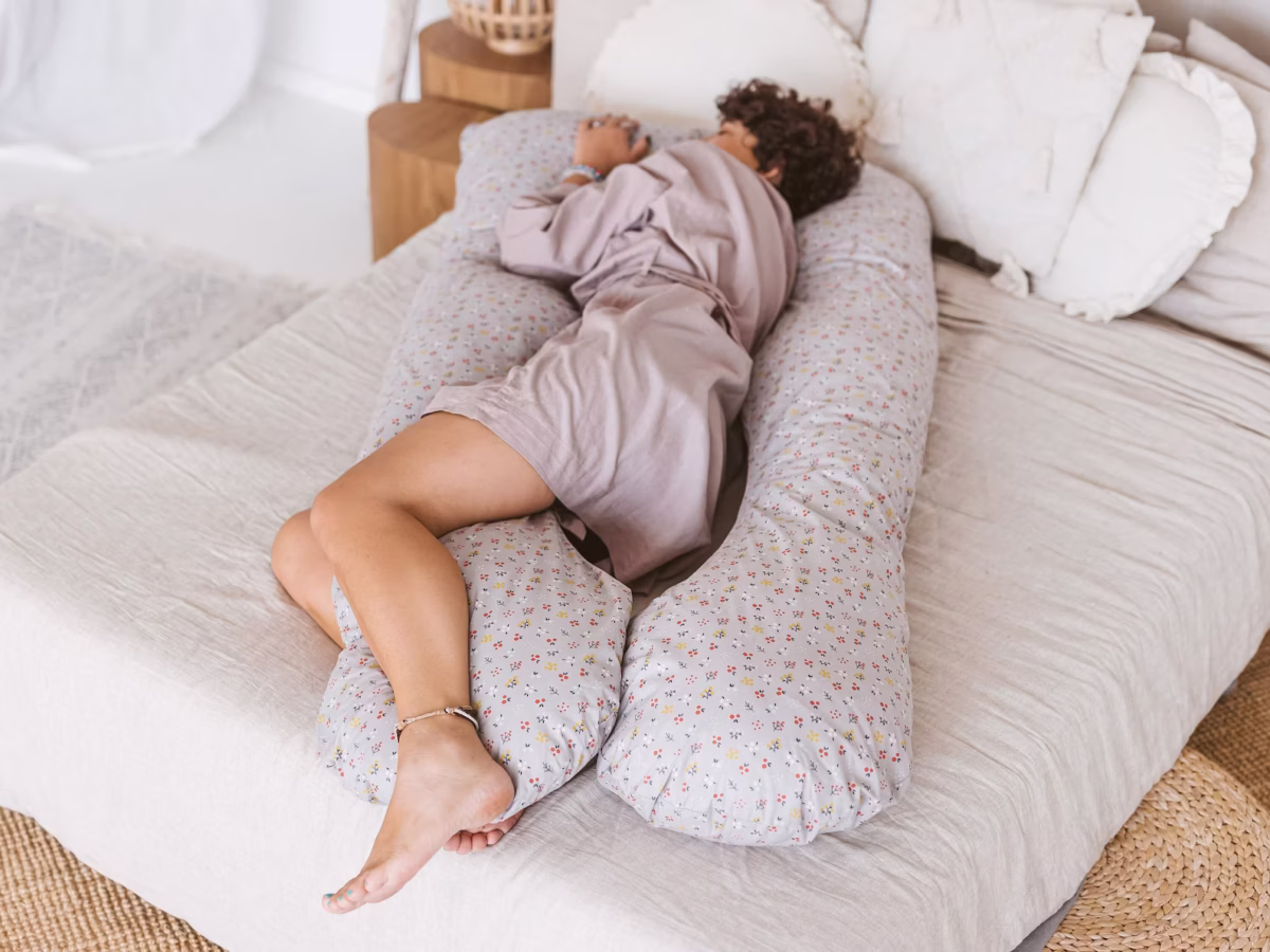 The photo shows a person in a purple robe sleeping on a bed while comfortably nested inside a large, U-shaped floral patterned body pillow.