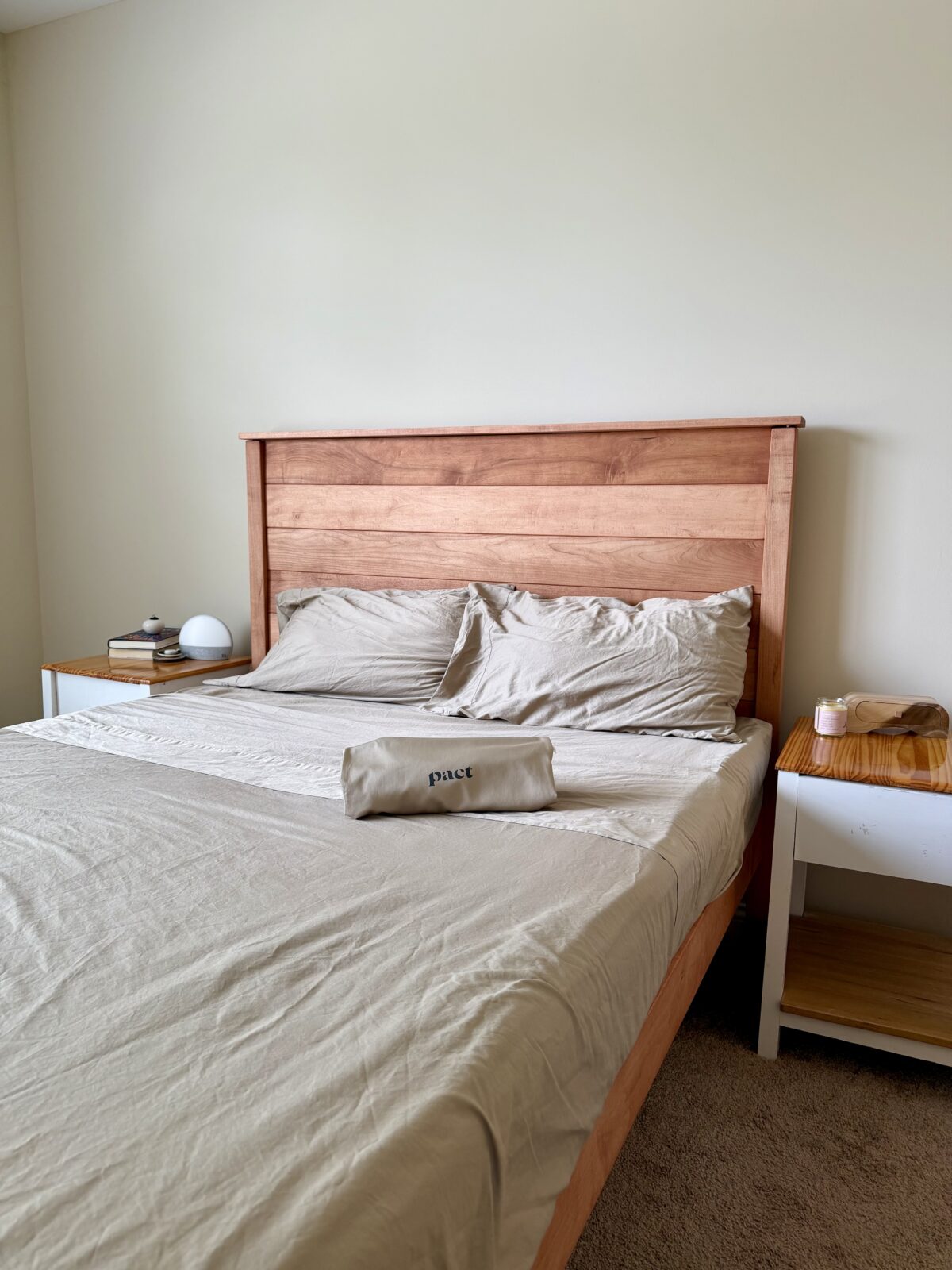 A neatly made bed with light beige sheets and two pillows, a wooden headboard, a nightstand with books and a lamp on the left, a tray on the right, and a beige pouch labeled pact placed on the bed.