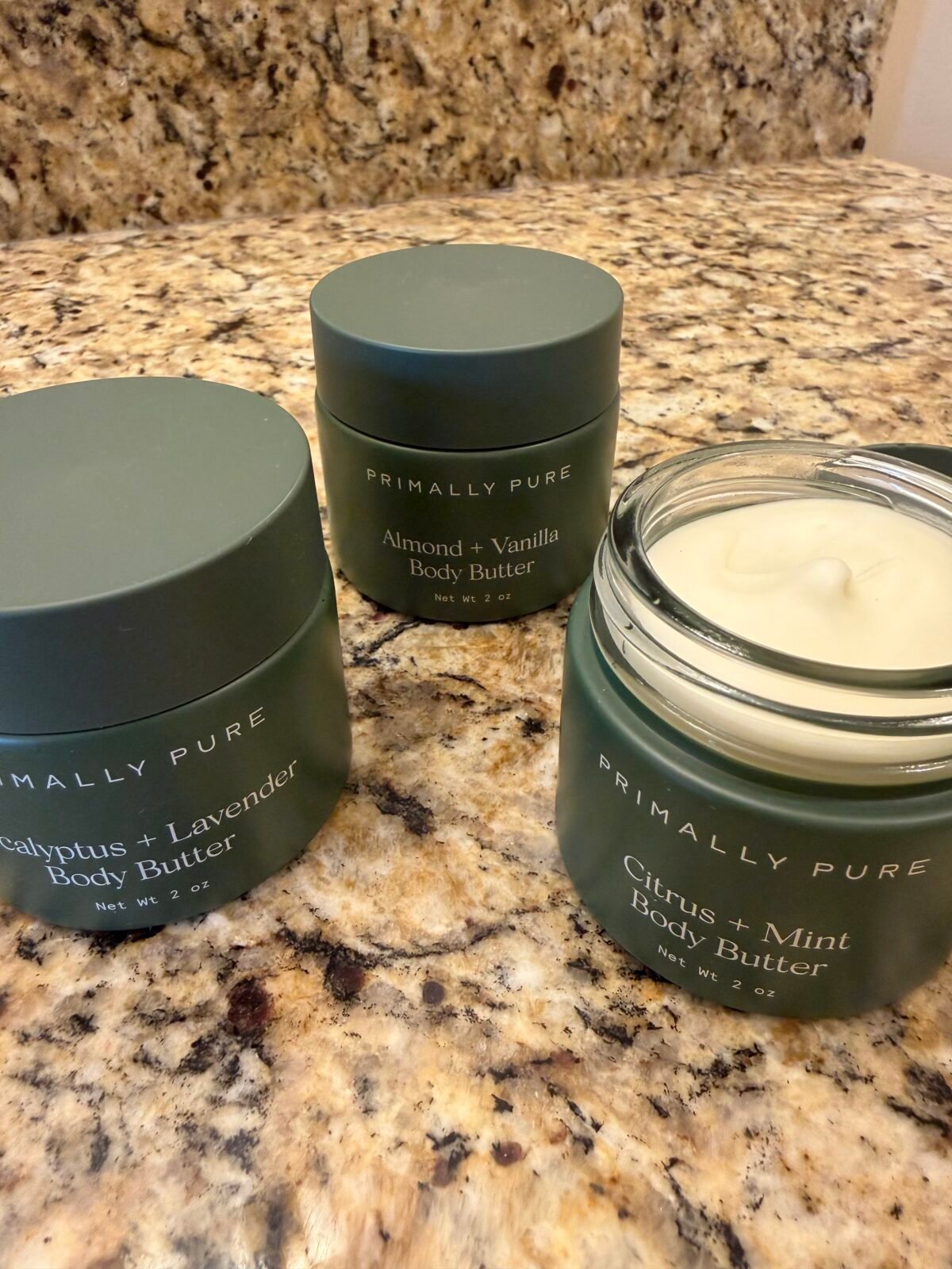 Three Primally Pure body butter jars in dark green containers on granite counter, showing Eucalyptus + Lavender, Almond + Vanilla, and Citrus + Mint varieties
