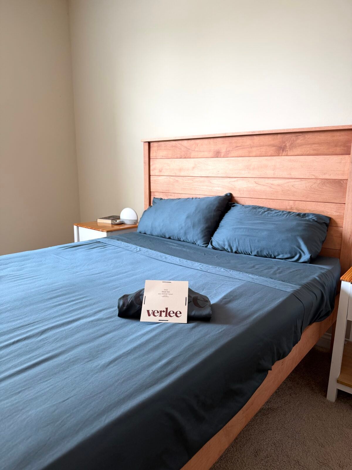 A neatly made bed with dark blue sheets and two pillows, a wooden headboard, and a card with the word verlee placed with black fabric on the bed. Nightstands with small items are on either side.