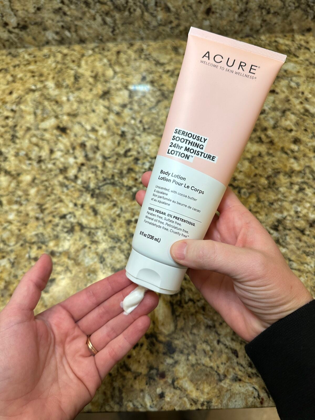 two hands holding acure's seriously soothing lotion with a bathroom counter in the background. One hand is squeezing some lotion out onto the other hand.