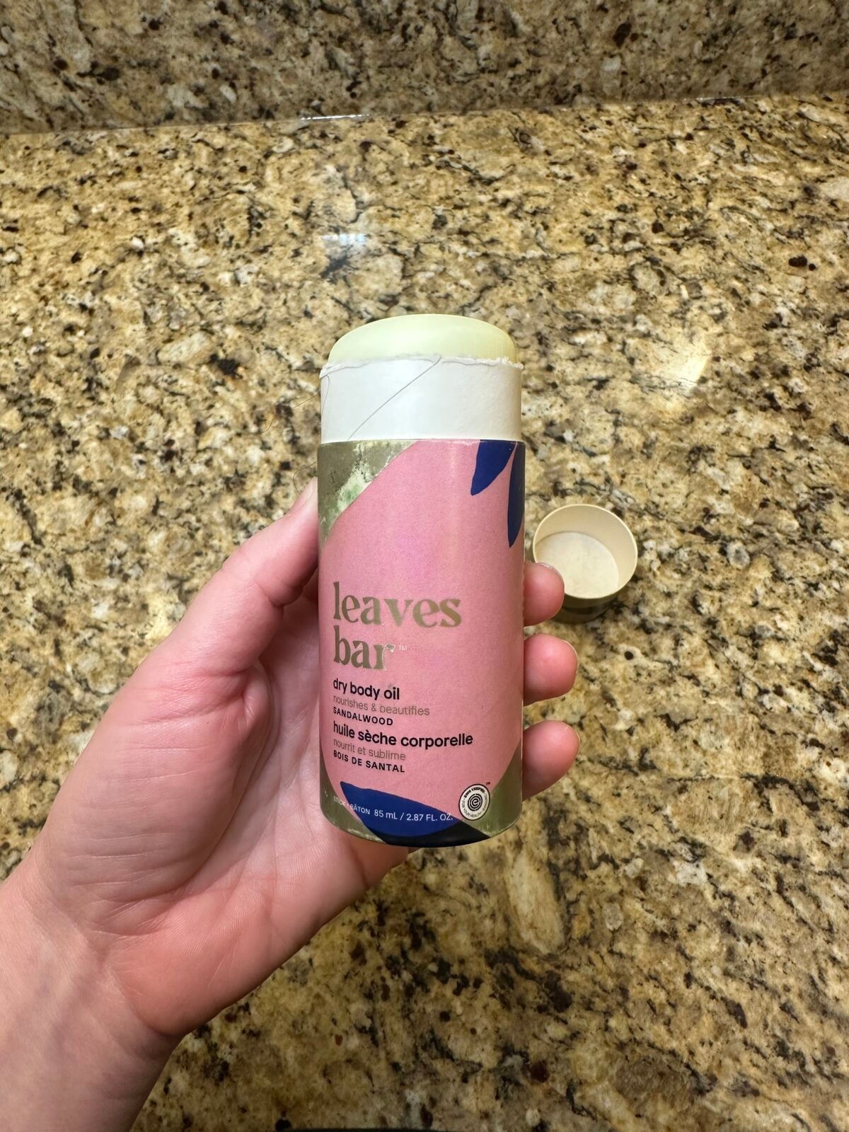 The photo shows a hand holding a pink and gold tube of Attitude "leaves bar" sandalwood dry body oil over a speckled granite countertop.