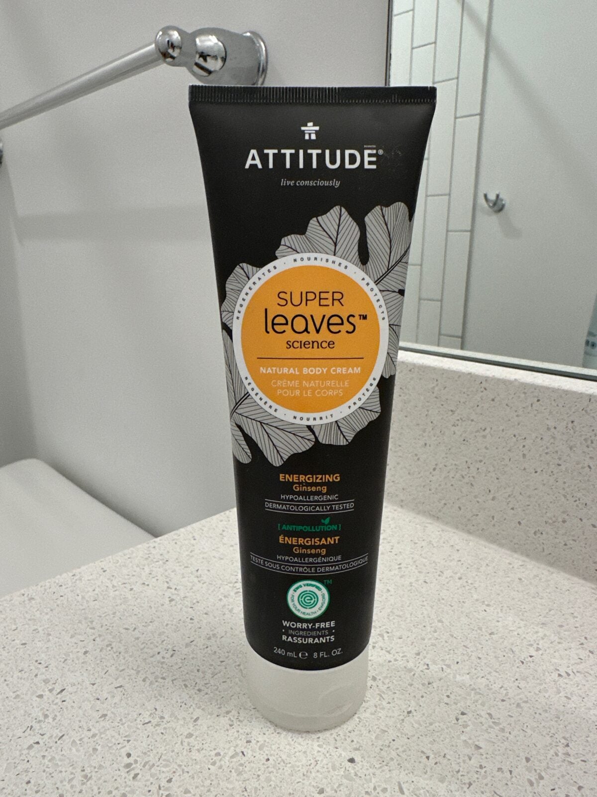 The photo shows an 8 fl oz black tube of Attitude "Super Leaves Science" energizing natural body cream with ginseng sitting on a white speckled bathroom countertop.