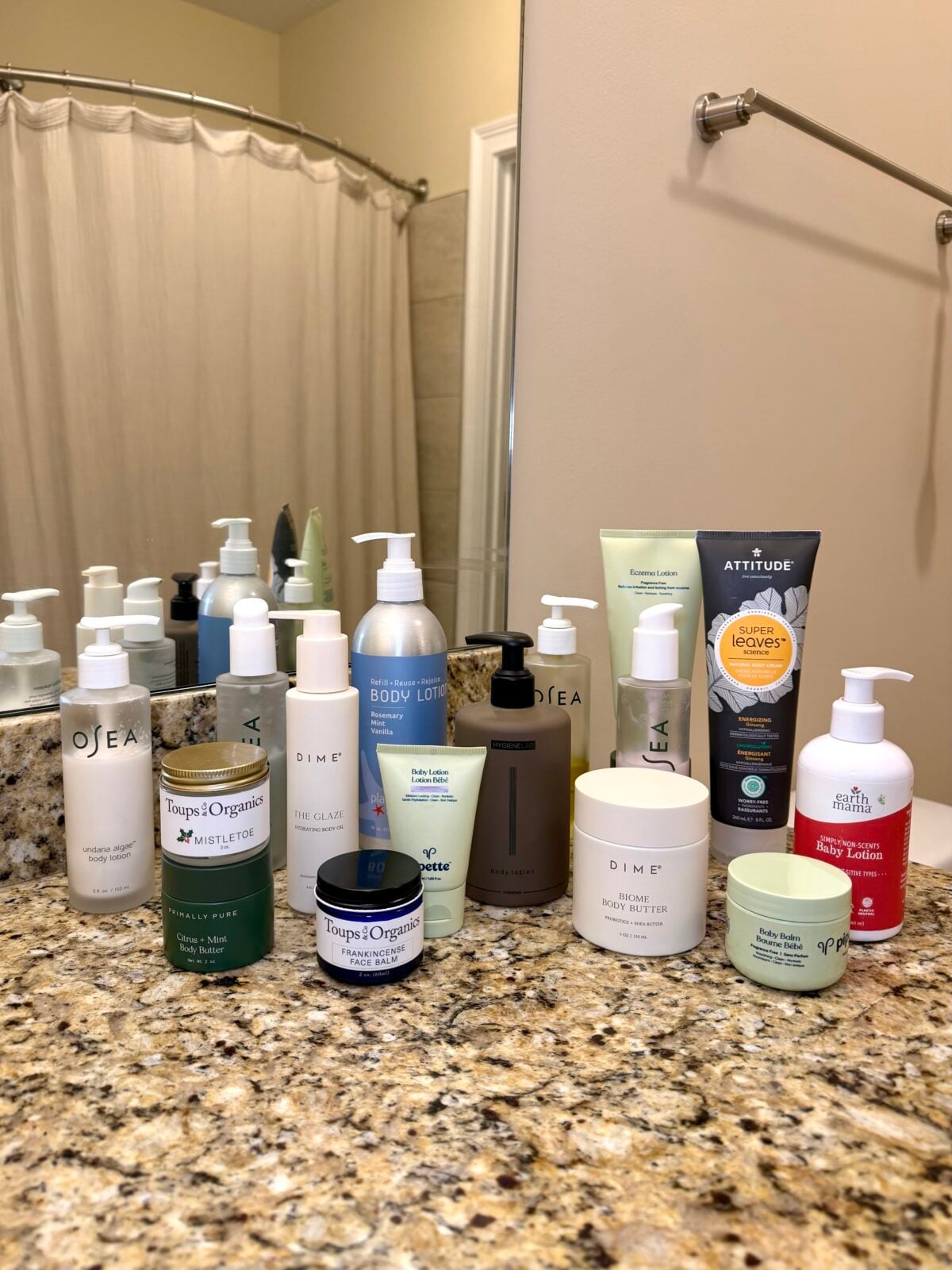 Assortment of natural body care products including brands like Osea, Dime, Attitude, and Earth Mama arranged on granite bathroom countertop
