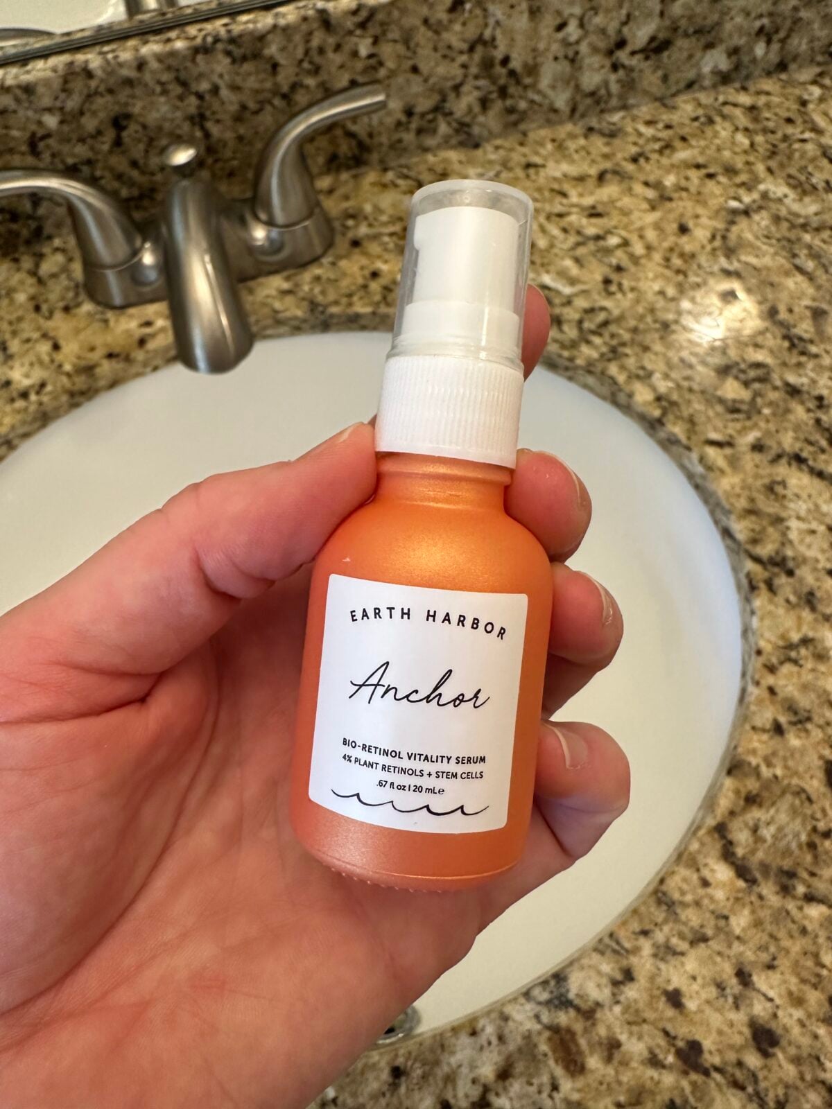 A hand holds a small, orange frosted glass pump bottle of Earth Harbor "Anchor" Bio-Retinol Vitality Serum over a white bathroom sink.