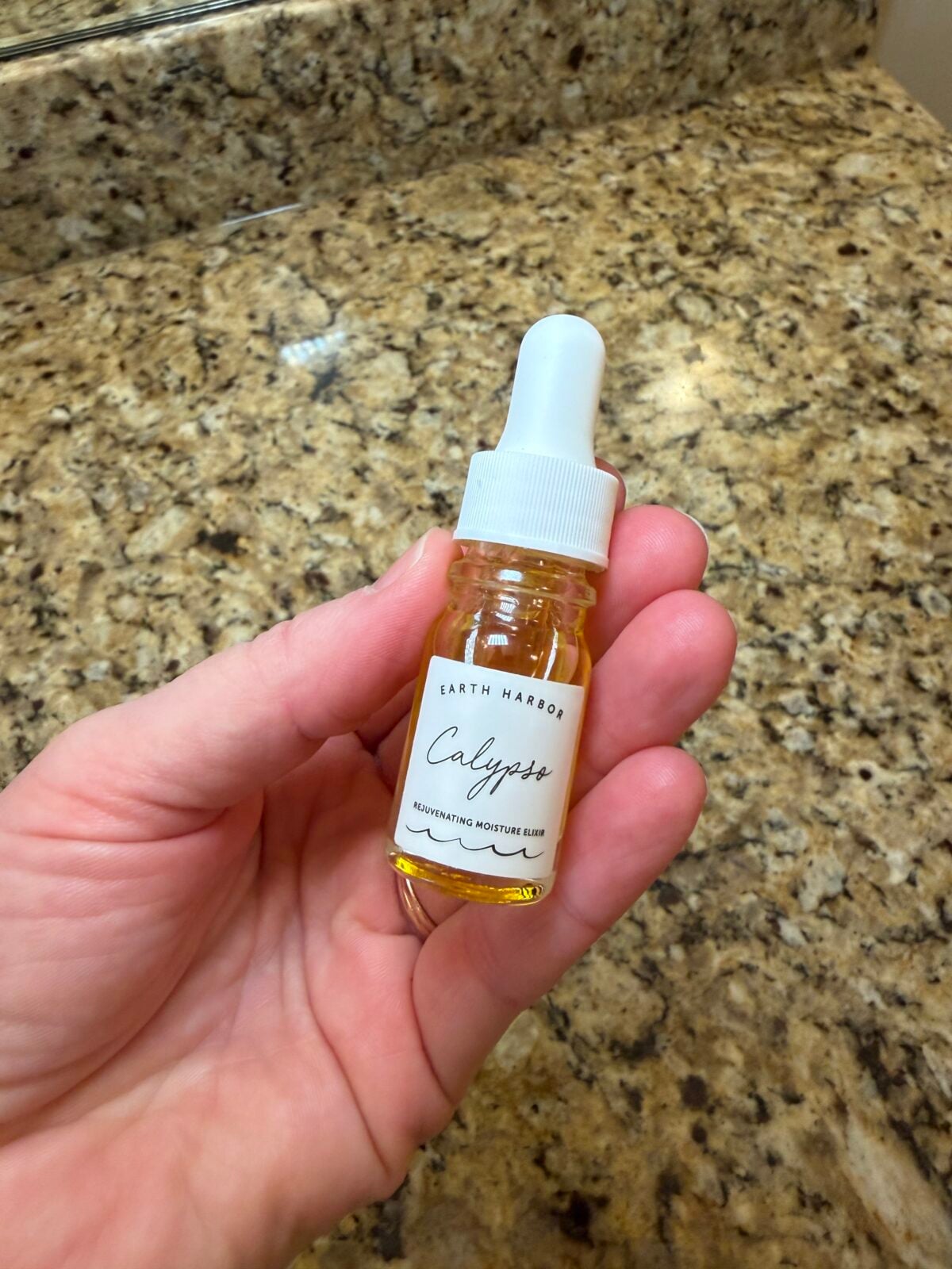 A hand holds a small glass dropper bottle of Earth Harbor "Calypso" Rejuvenating Moisture Elixir, which contains a golden-colored oil, over a speckled granite countertop.