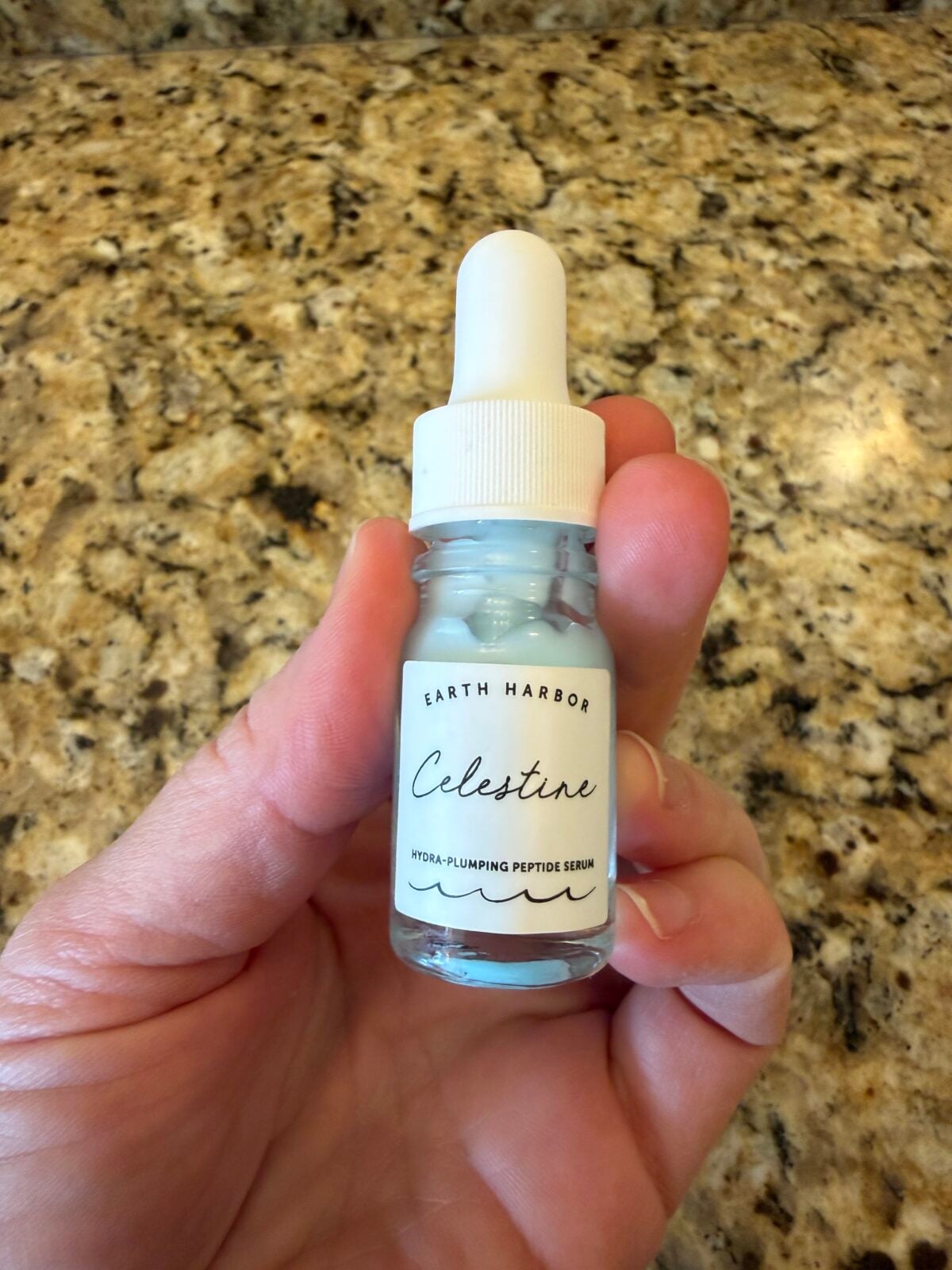 A hand holds a small glass dropper bottle of Earth Harbor "Celestine" Hydra-Plumping Peptide Serum, which contains a light blue liquid, against a background of a speckled granite countertop.
