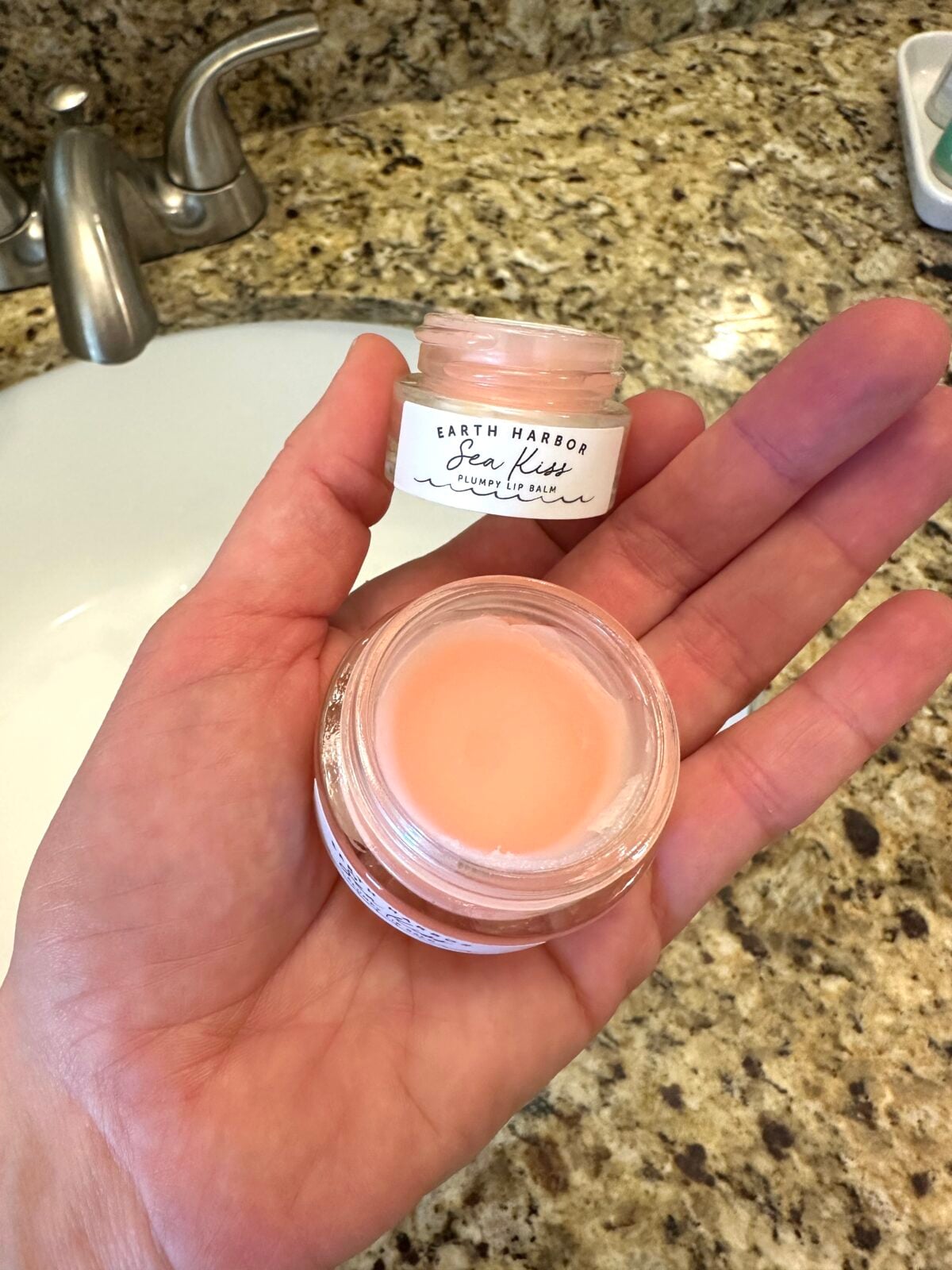 Hand holding open jar of Earth Harbor Sea Kiss Plumpy Lip Balm showing peach-colored product over bathroom sink