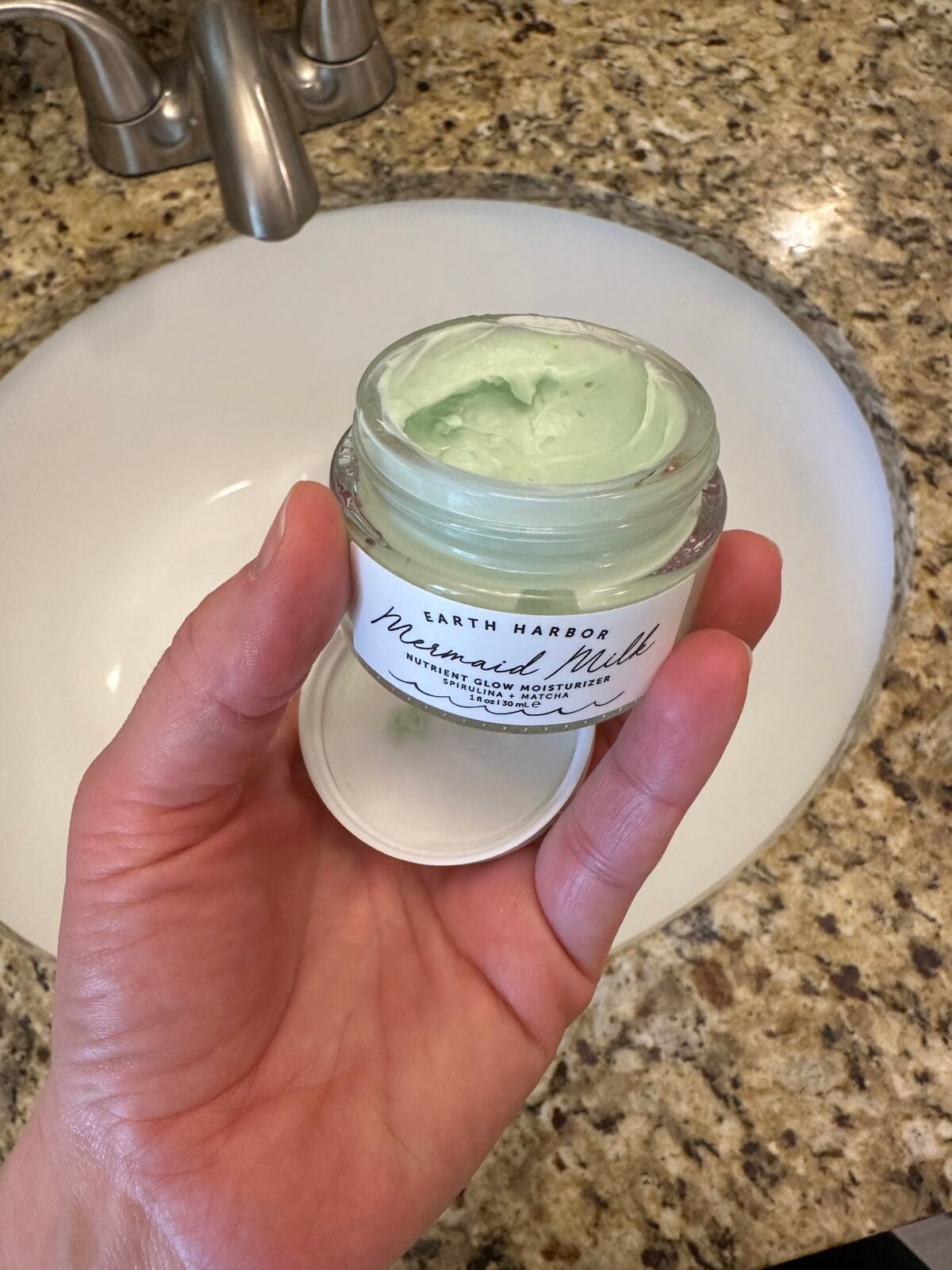 A hand holds an open glass jar of Earth Harbor "Mermaid Milk" Nutrient Glow Moisturizer, revealing the pale green cream inside, over a white bathroom sink.