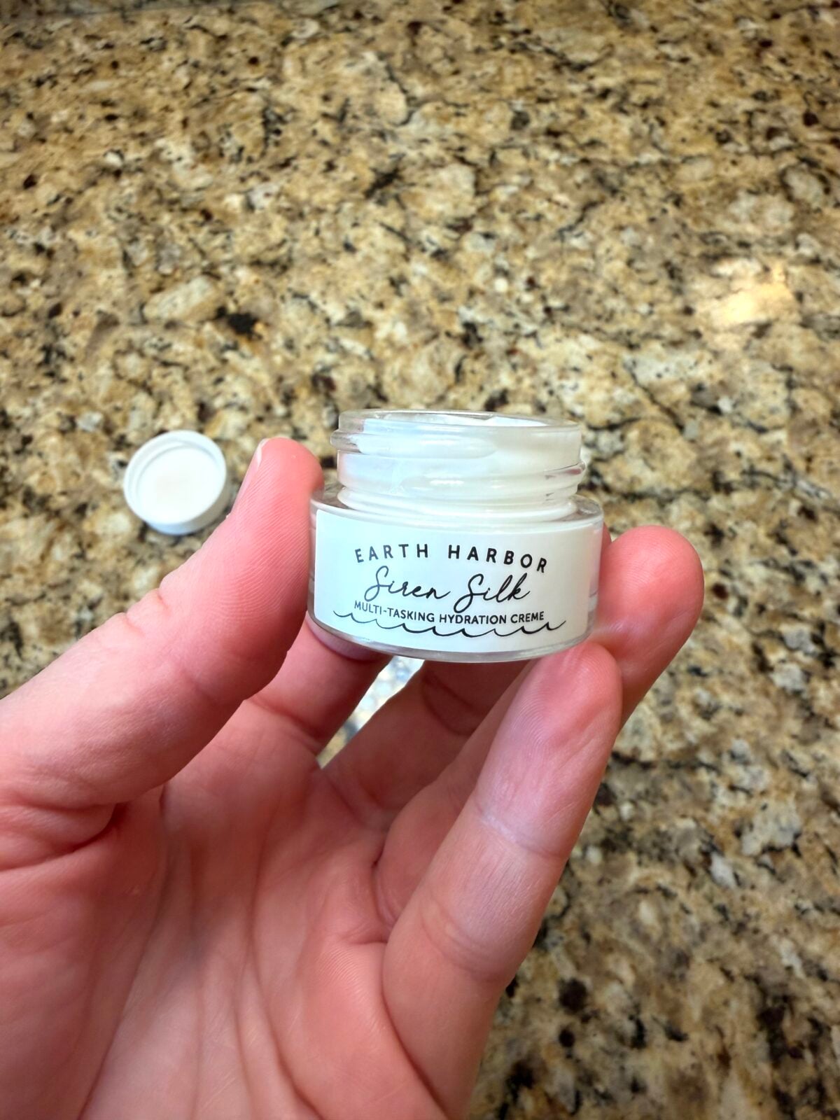 The photo shows a hand holding an open glass jar of Earth Harbor "Siren Silk" Multi-Tasking Hydration Creme, revealing the white cream inside, over a speckled granite countertop.