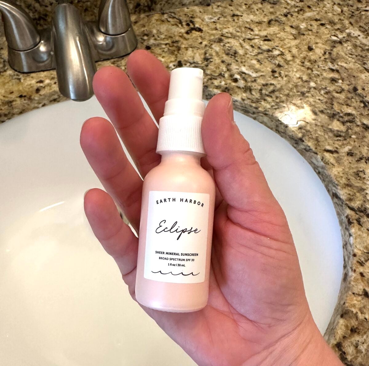 A hand holds a pale pink bottle of Earth Harbor "Eclipse" Sheer Mineral Sunscreen, which features a broad spectrum SPF 30 rating, over a white bathroom sink.