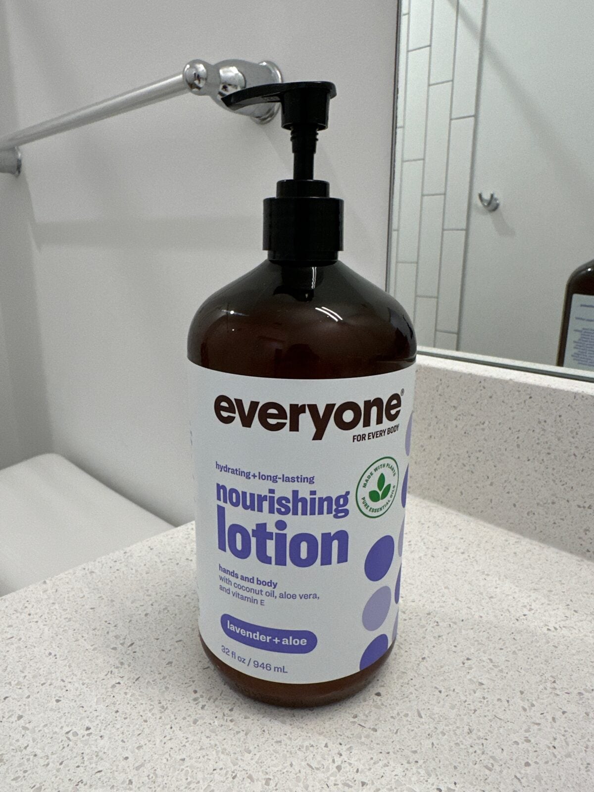 The photo shows a large 32 fl oz amber pump bottle of Everyone "Nourishing Lotion" in Lavender + Aloe scent sitting on a white speckled bathroom countertop.
