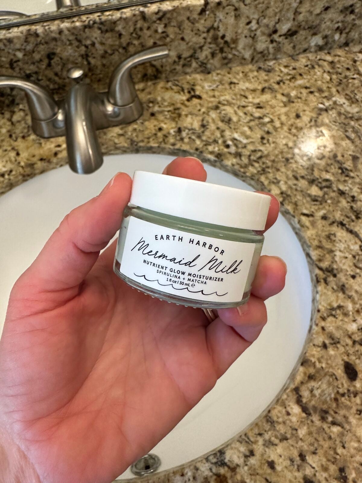 A hand holds a small glass jar of Earth Harbor "Mermaid Milk" Nutrient Glow Moisturizer over a white bathroom sink with a granite countertop.