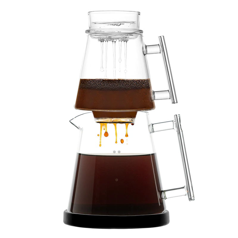 The photo shows a clear glass pour-over coffee brewer where water is dripping through coffee grounds in an upper chamber into a lower carafe filled with brewed coffee.