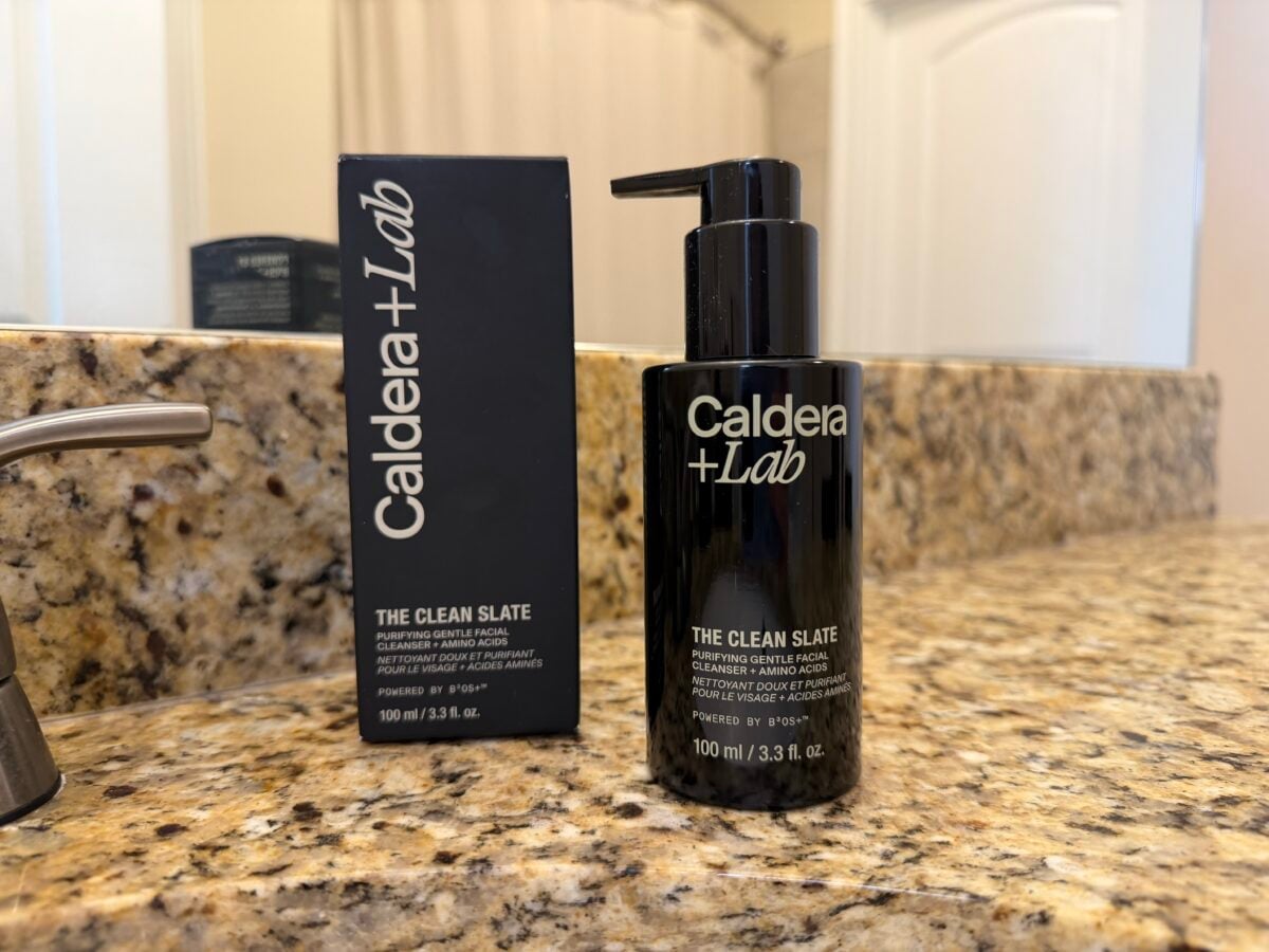 A black bottle labeled Caldera + Lab The Clean Slate with a pump sits on a granite bathroom counter next to its matching black box packaging. A faucet and mirror are visible in the background.