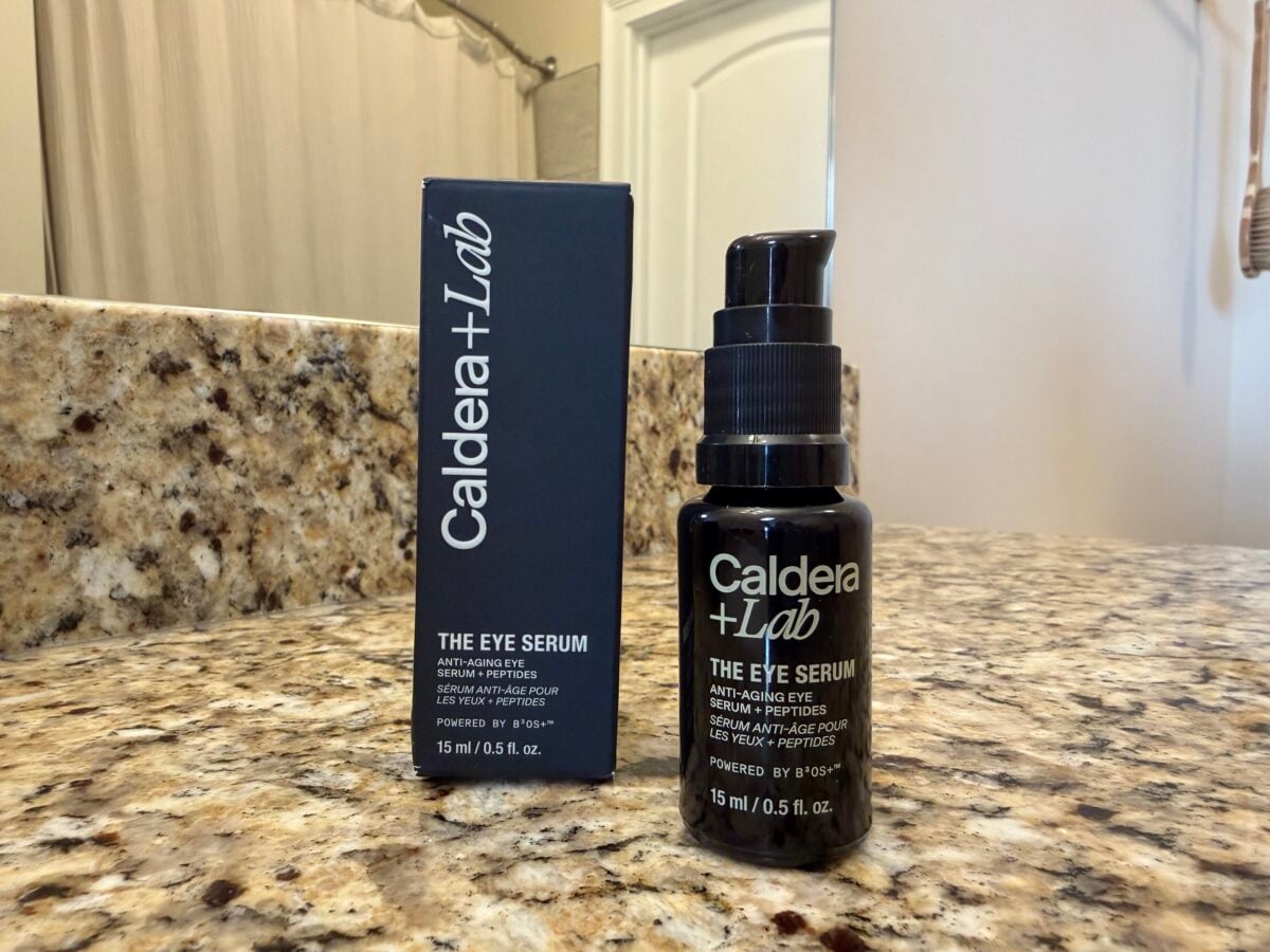 A bottle and box of Caldera + Lab The Eye Serum sit on a granite bathroom countertop. The box is upright to the left, and the bottle with a pump is on the right in front of a bathroom mirror.