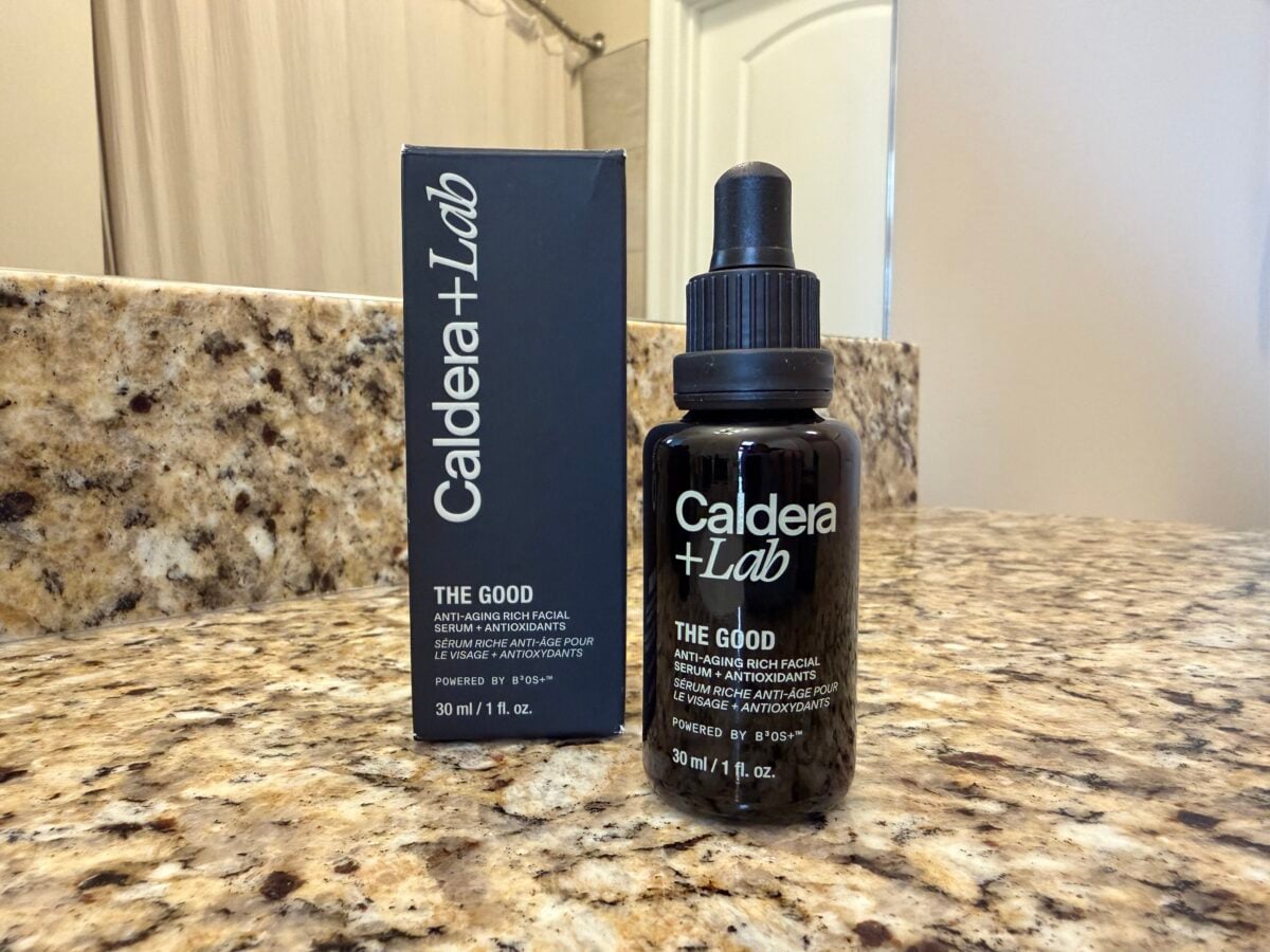 A bottle and box of Caldera + Lab The Good facial serum sit on a granite bathroom countertop. The bottle has a dropper cap and both items display product information in white text.