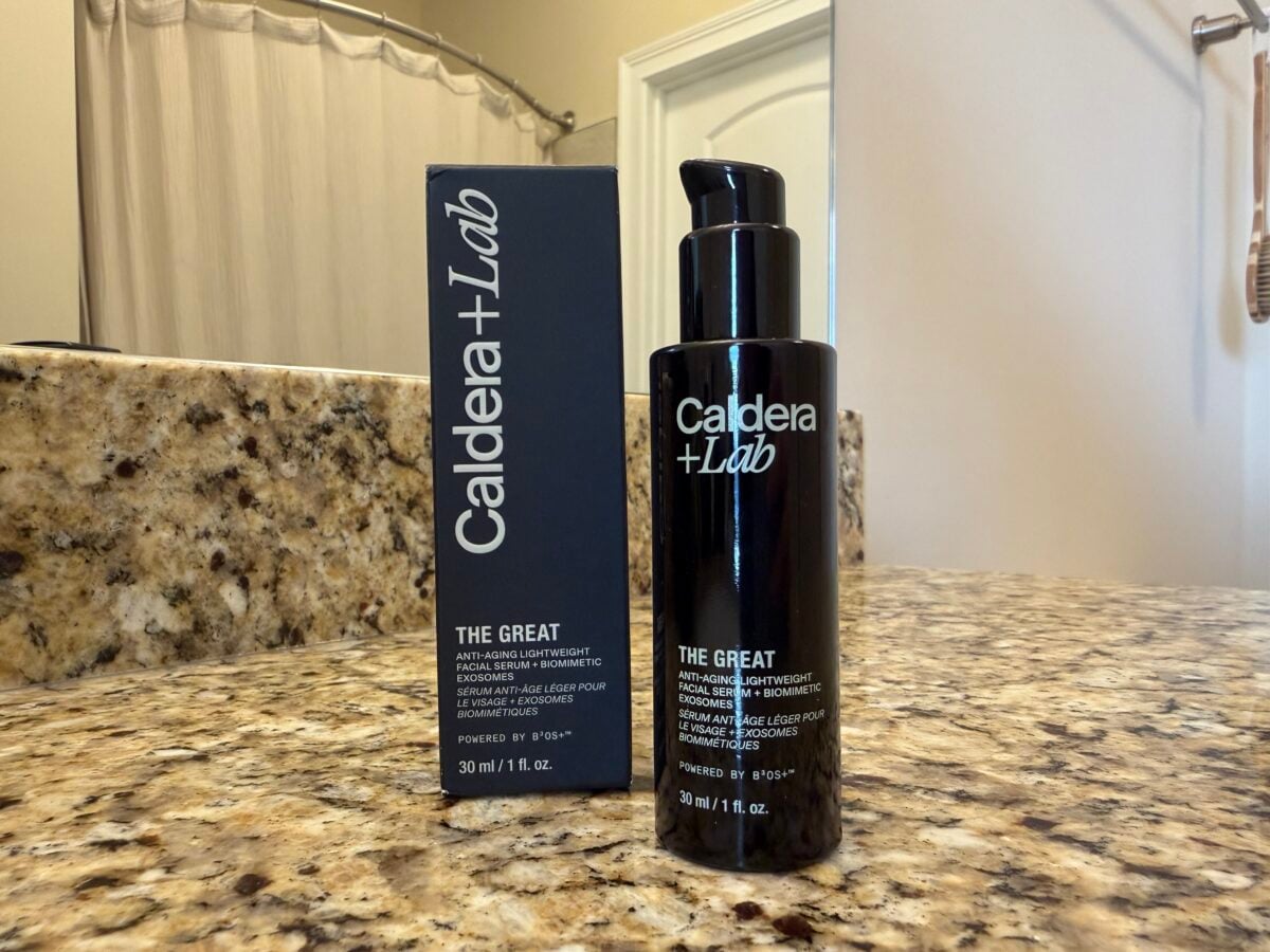 A black bottle of Caldera + Lab The Great serum with a pump dispenser is placed next to its matching box on a bathroom counter with a granite surface. A mirror, sink, and towel are in the background.