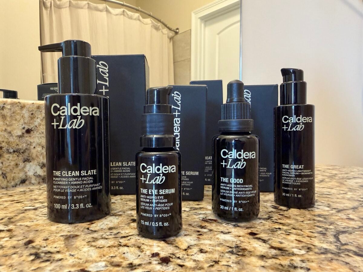 Four Caldera + Lab skincare products, including The Clean Slate, The Base Layer, The Good, and The Great, are displayed on a bathroom countertop with product boxes and a shower curtain in the background.