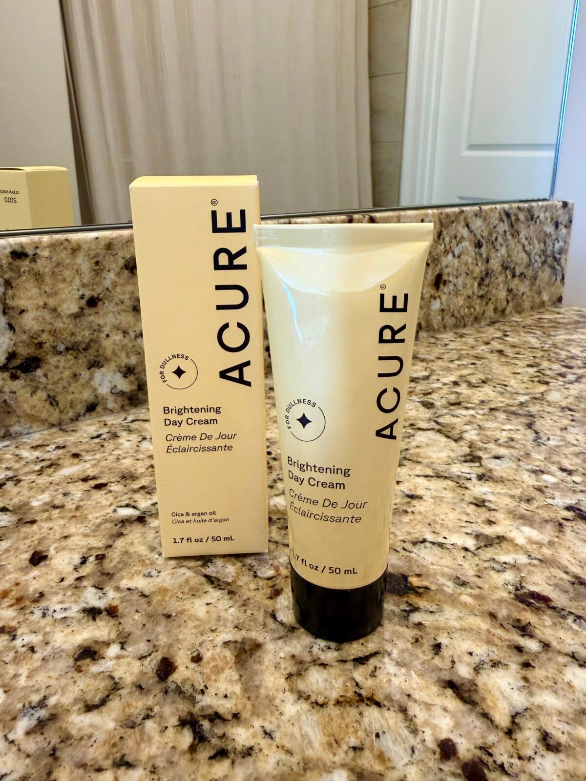 A tube of Acure Brightening Day Cream is placed on a granite bathroom counter in front of its matching box. Both items are labeled 1.7 fl oz / 50 ml. A mirror and part of a sink are visible in the background.