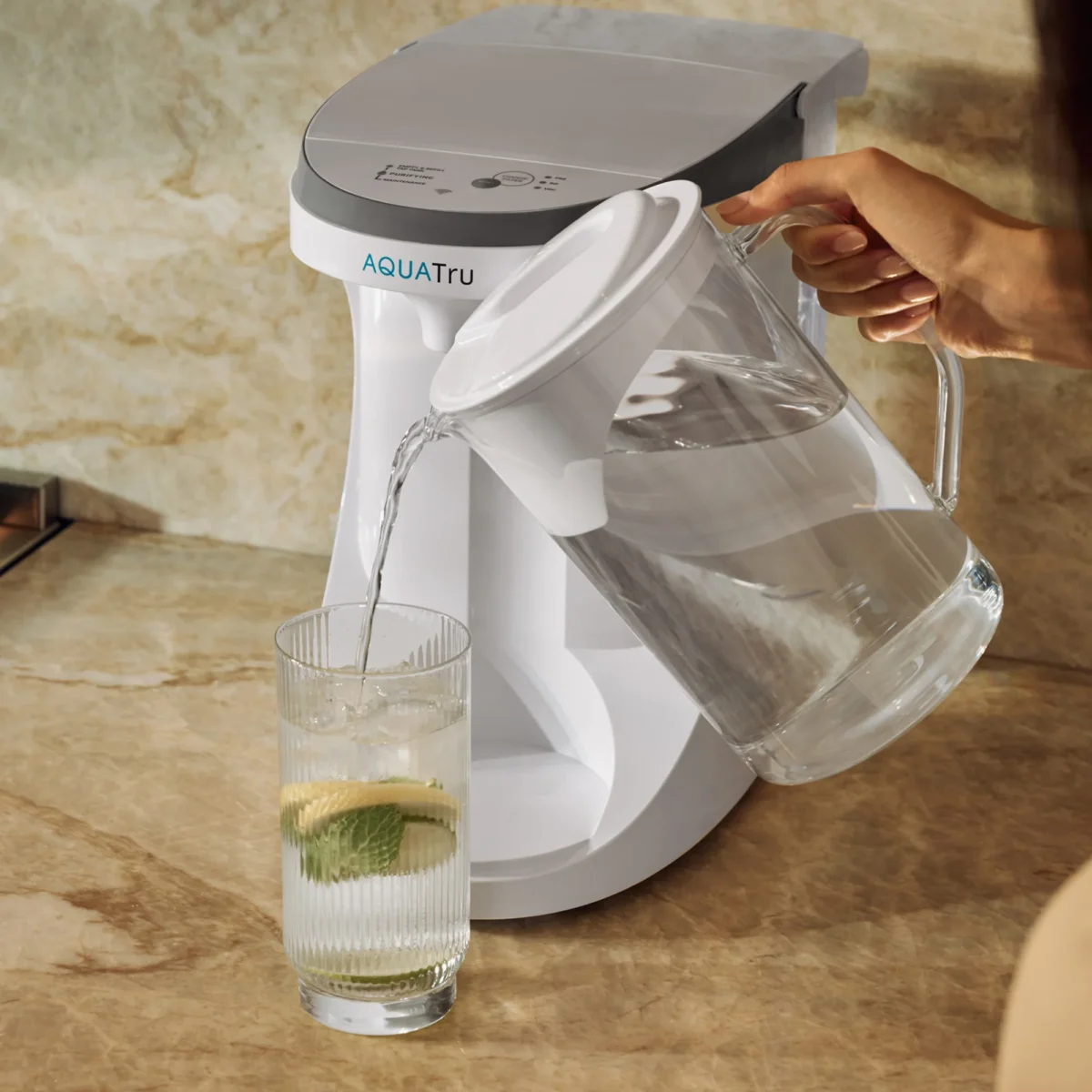 A person's hand is shown pouring filtered water from a clear pitcher into a glass with lemon and mint, situated next to an AquaTru countertop reverse osmosis system.