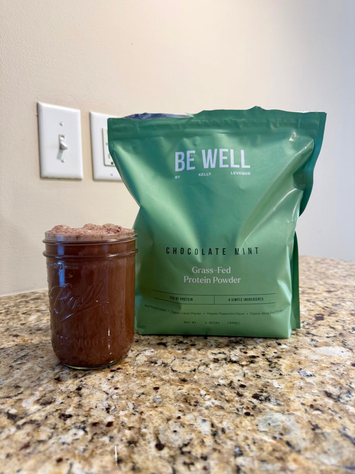 A green bag labeled BE WELL Chocolate Mint Grass-Fed Protein Powder stands on a granite countertop next to a glass jar filled with a chocolate-colored drink. Two light switches are visible on the wall behind.