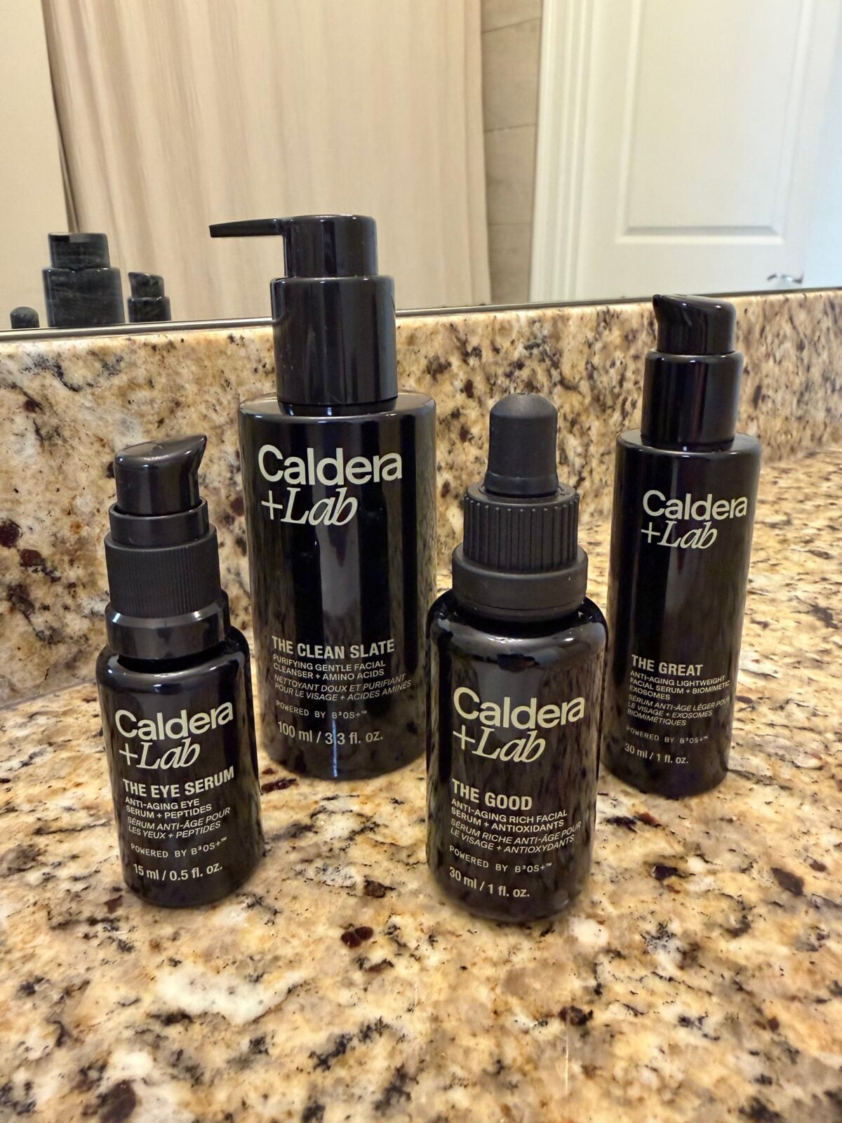 Four Caldera + Lab skincare products—The Eye Serum, The Clean Slate, The Good, and The Great—are arranged on a speckled granite bathroom countertop in front of a mirror.