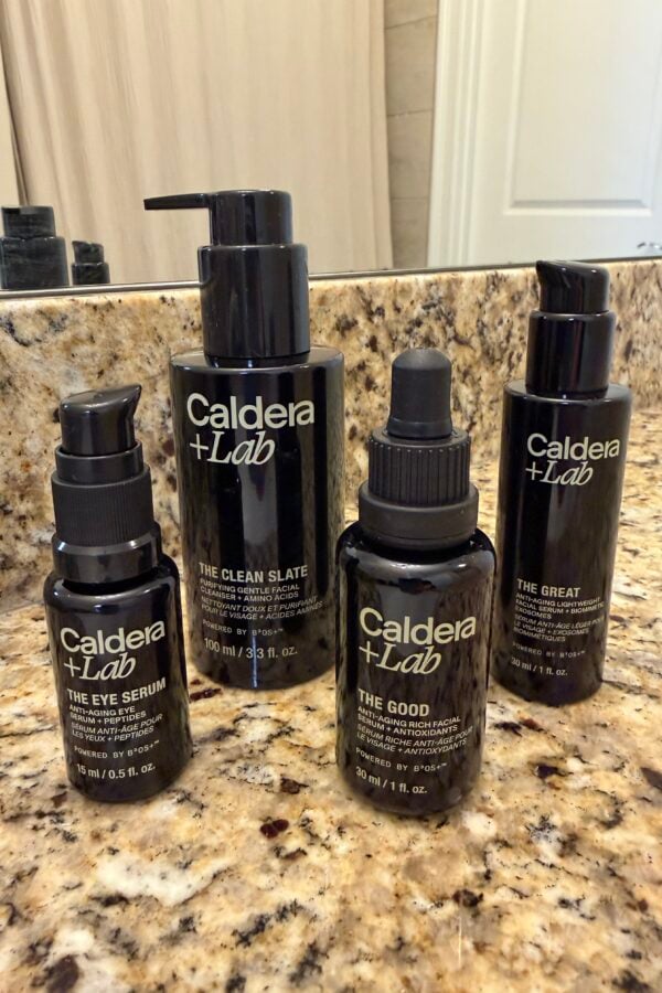 Four Caldera + Lab skincare products—The Eye Serum, The Clean Slate, The Good, and The Great—are arranged on a speckled granite bathroom countertop in front of a mirror.