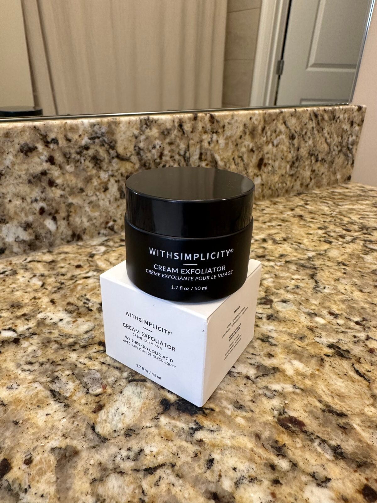 A black jar labeled &ldquo;WITHSIMPLICITY CREAM EXFOLIATOR&rdquo; sits on a granite countertop, with its white product box placed beneath it. A bathroom mirror, sink, and door are visible in the background.