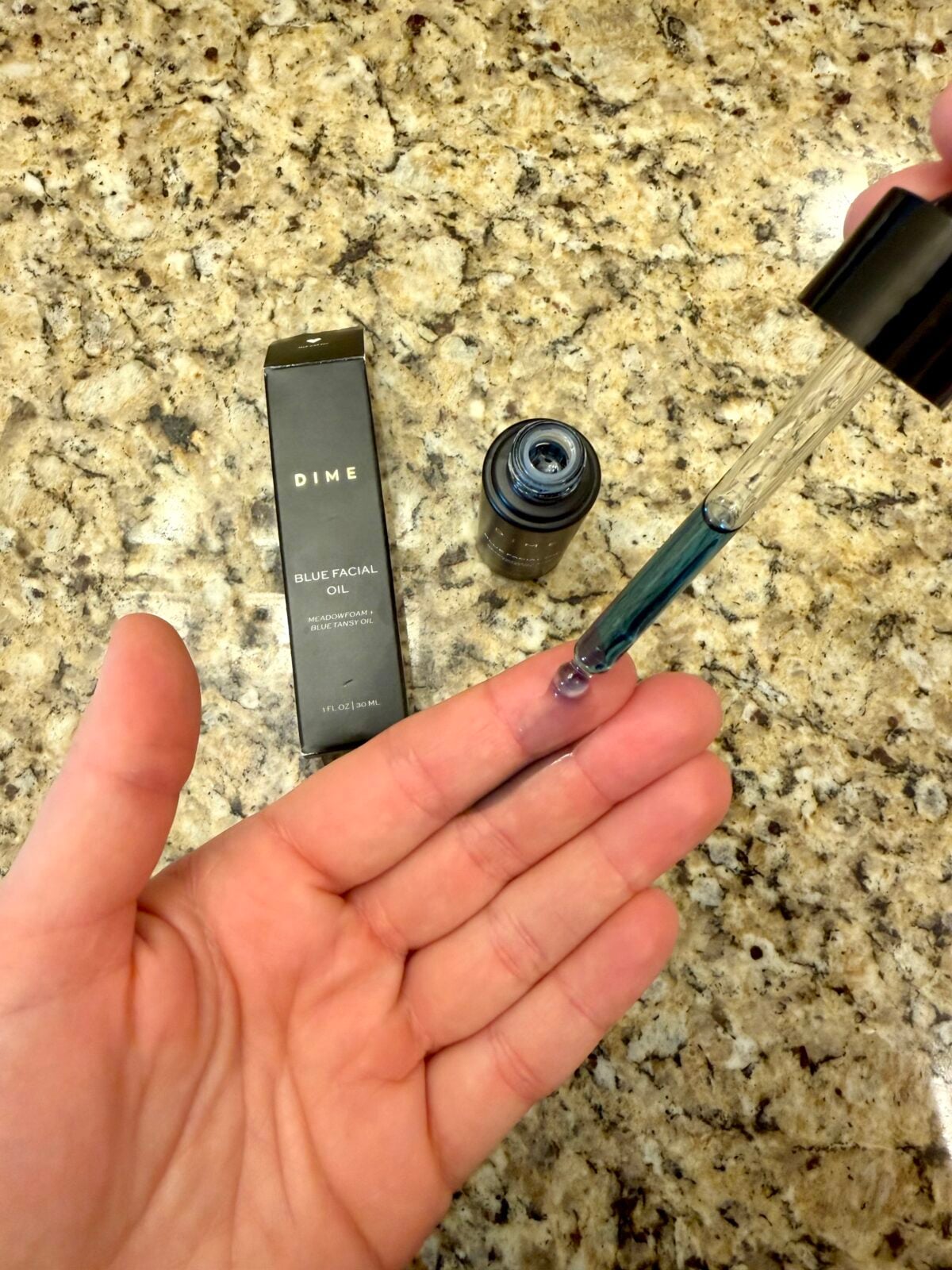 A hand holds a dropper with blue liquid above an open palm. A dark bottle labeled DIME Blue Facial Oil and its box are on a granite countertop.